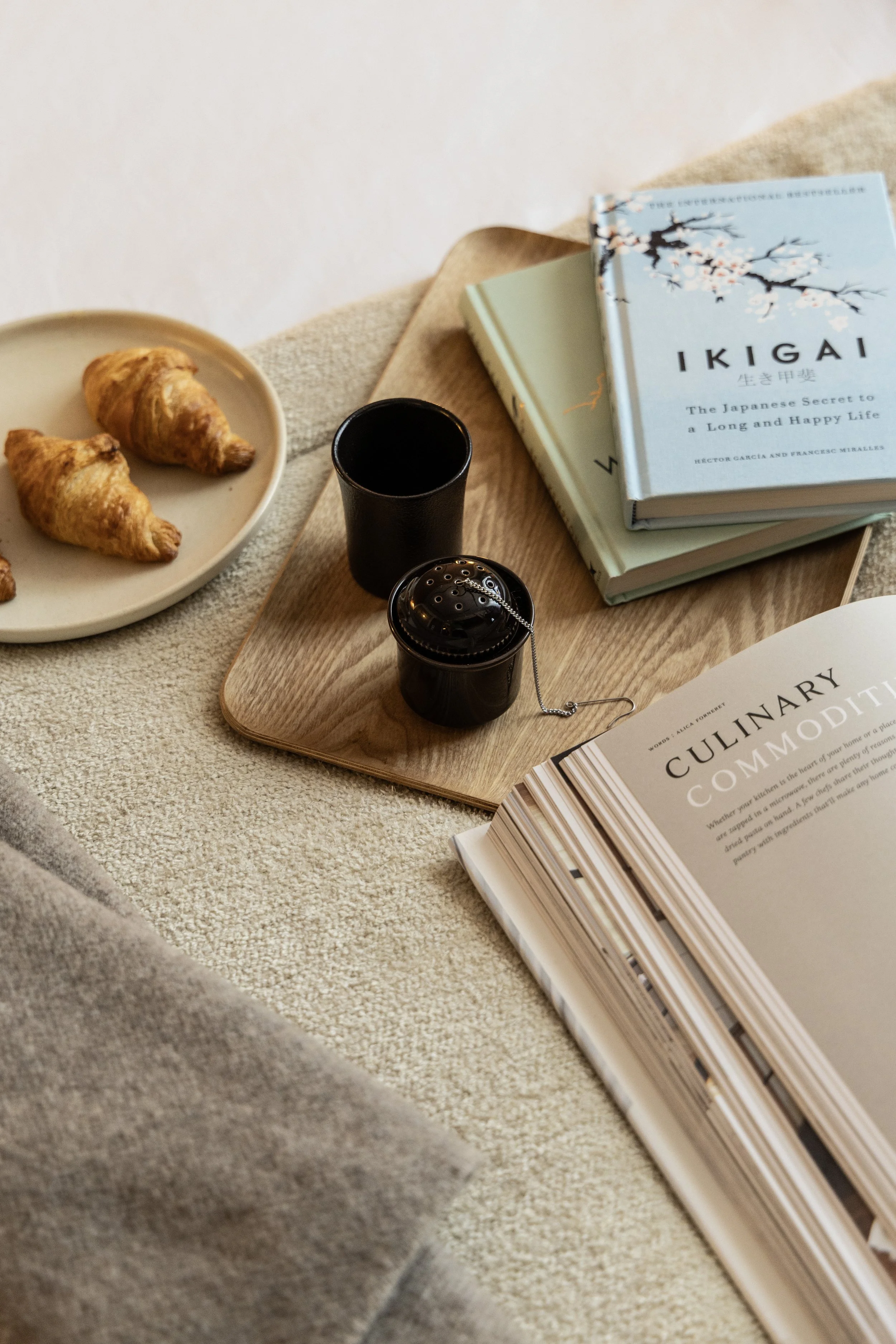A cozy table setup with a plate of three croissants, two black candles, a small black container with a chain, and several books, including one open cookbook titled 'Culinary.'