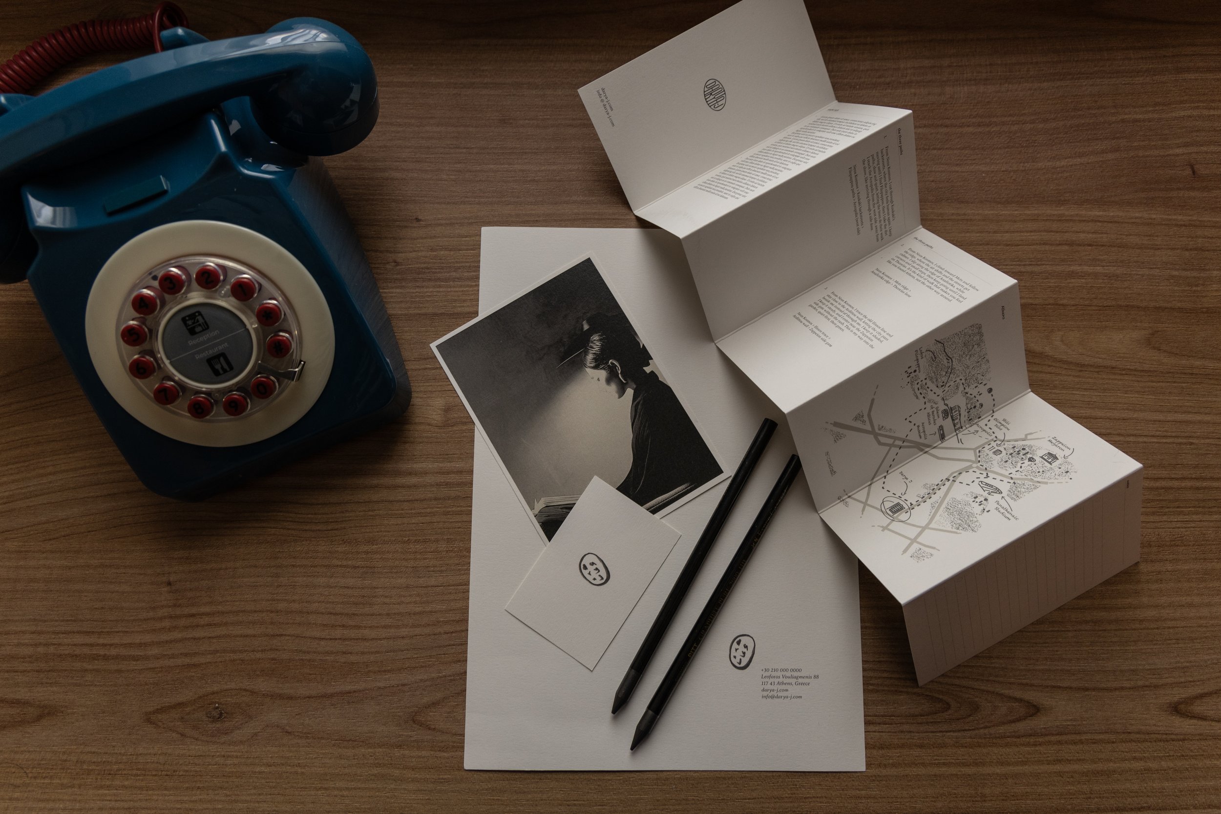 A vintage blue rotary phone on a wooden table, alongside open paper documents, photographs, two black pens, and small cards with smiley faces.