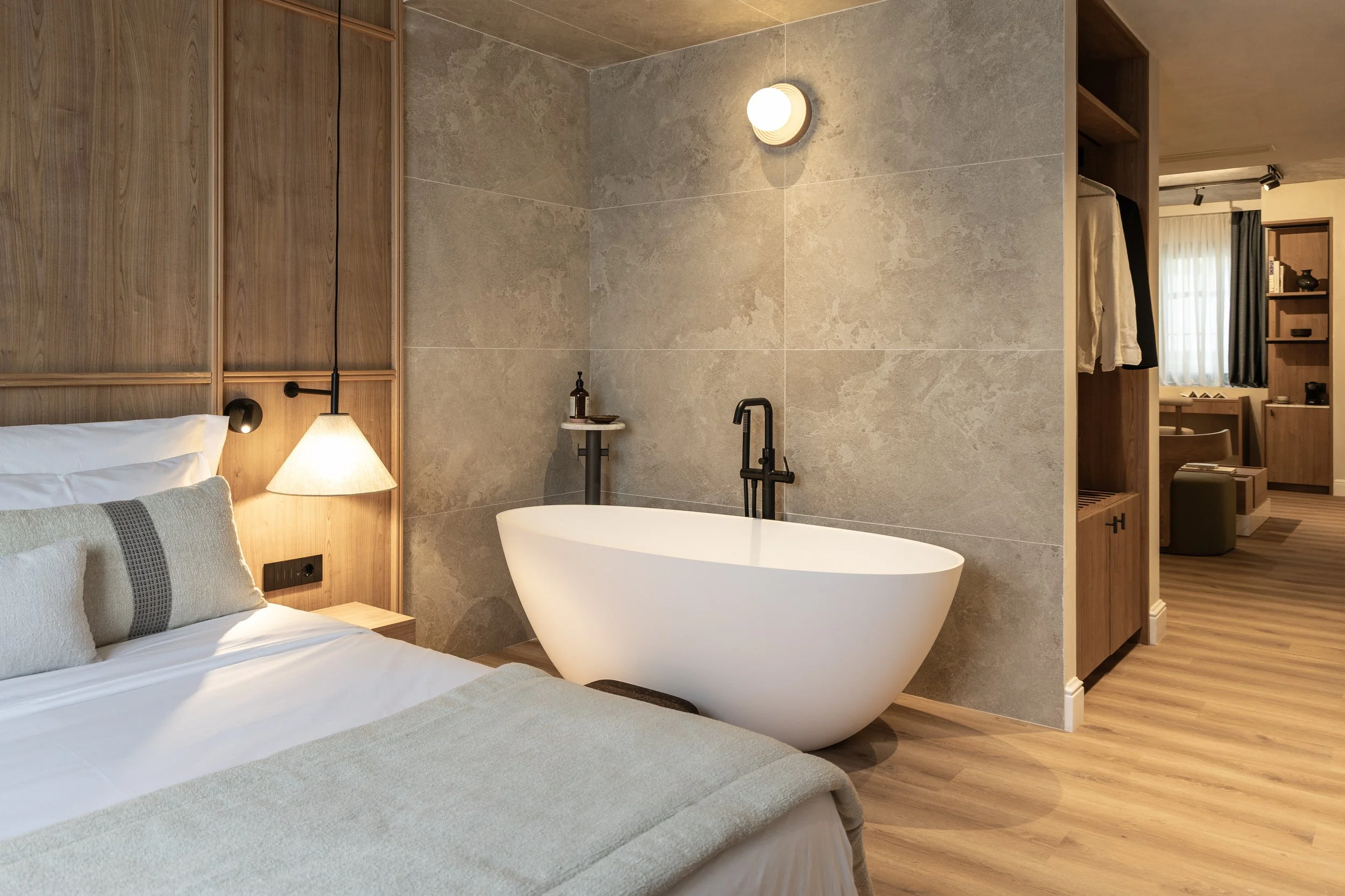 Hotel bedroom with a white bed and a standalone bathtub behind it, against a grey stone wall with a circular wall lamp, and part of a wooden wardrobe and sitting area in the background.