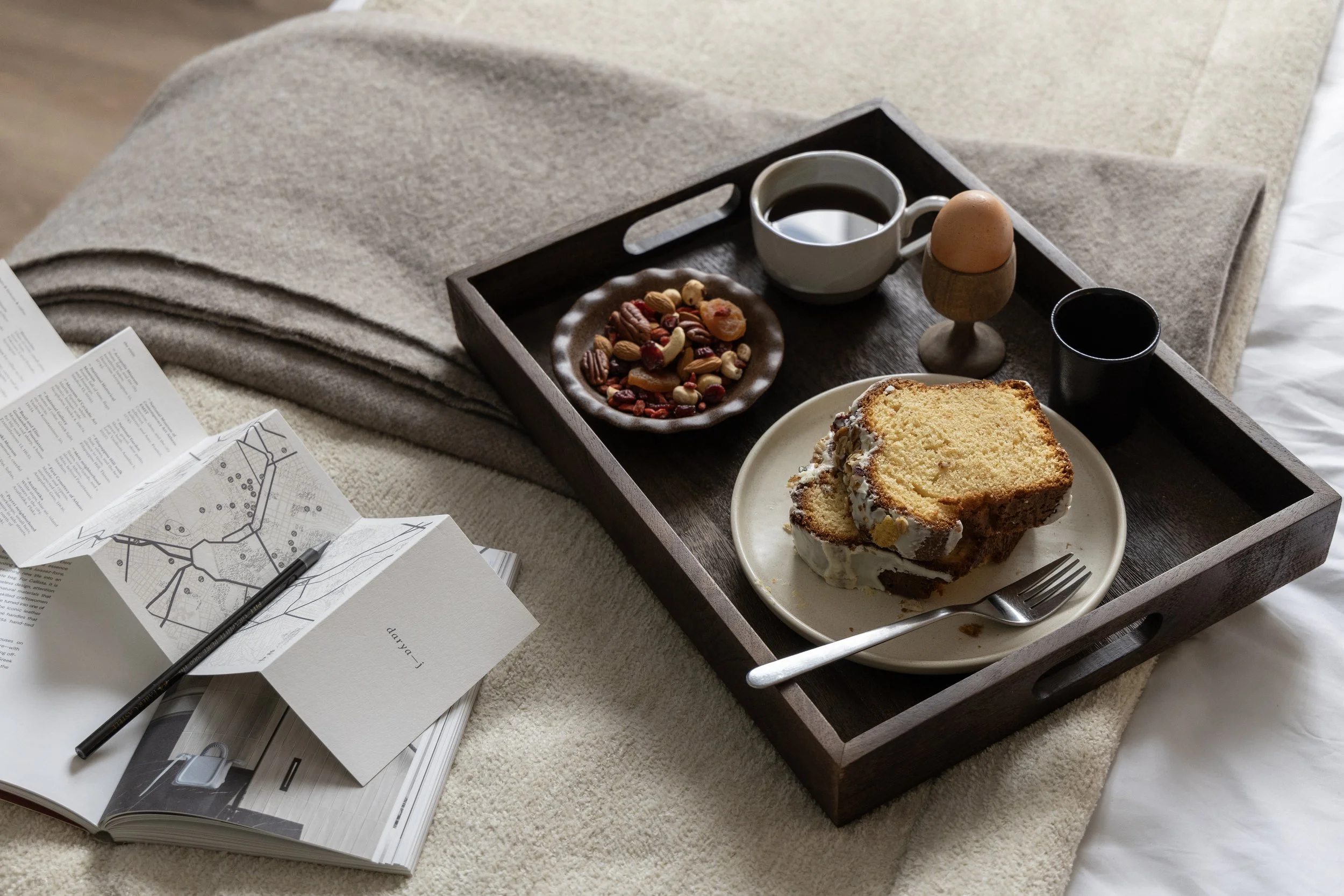 Tray with coffee, a soft-boiled egg in an egg holder, a small cup of black liquid, a plate with a slice of cake, a small plate of mixed nuts and dried fruit, and a mug of black coffee, placed on a bed with blankets and an open magazine.