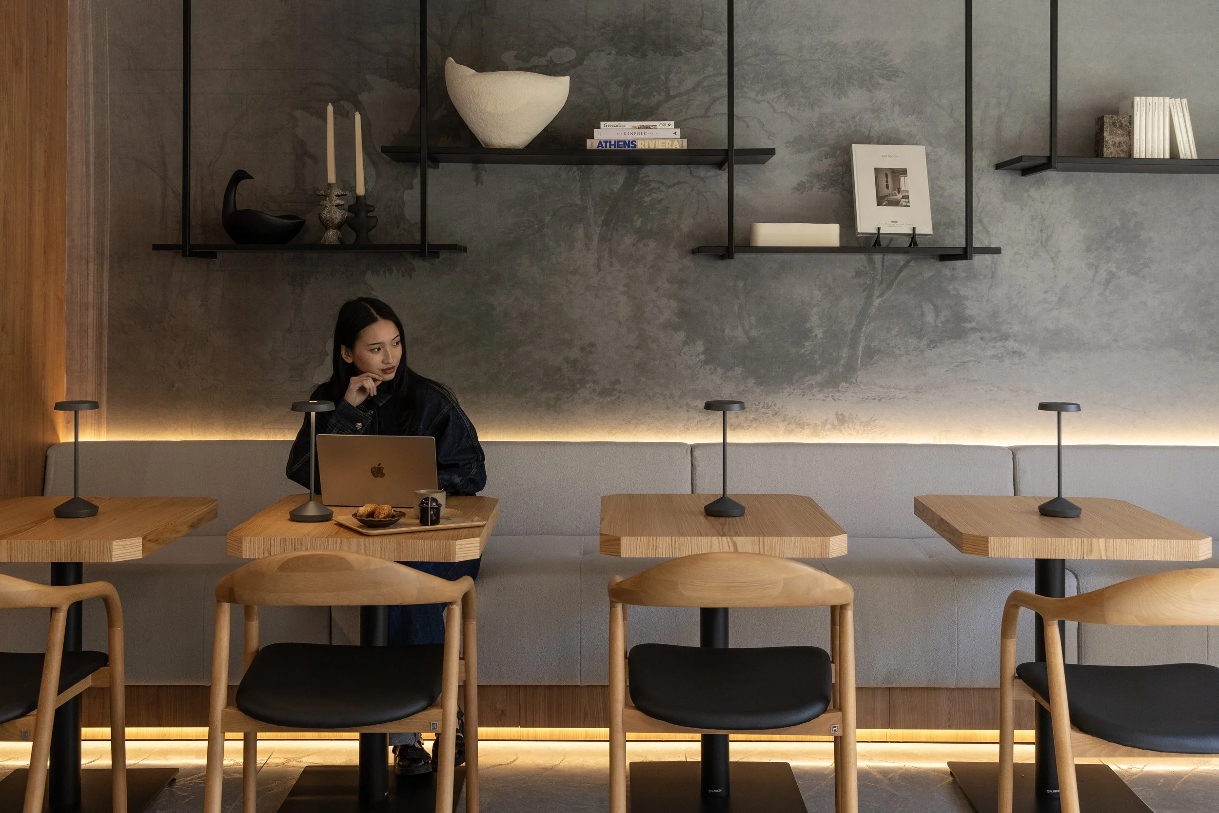 A young woman sitting at a wooden table with a laptop and snacks in a modern cafe with gray walls, black shelves, and wooden chairs.