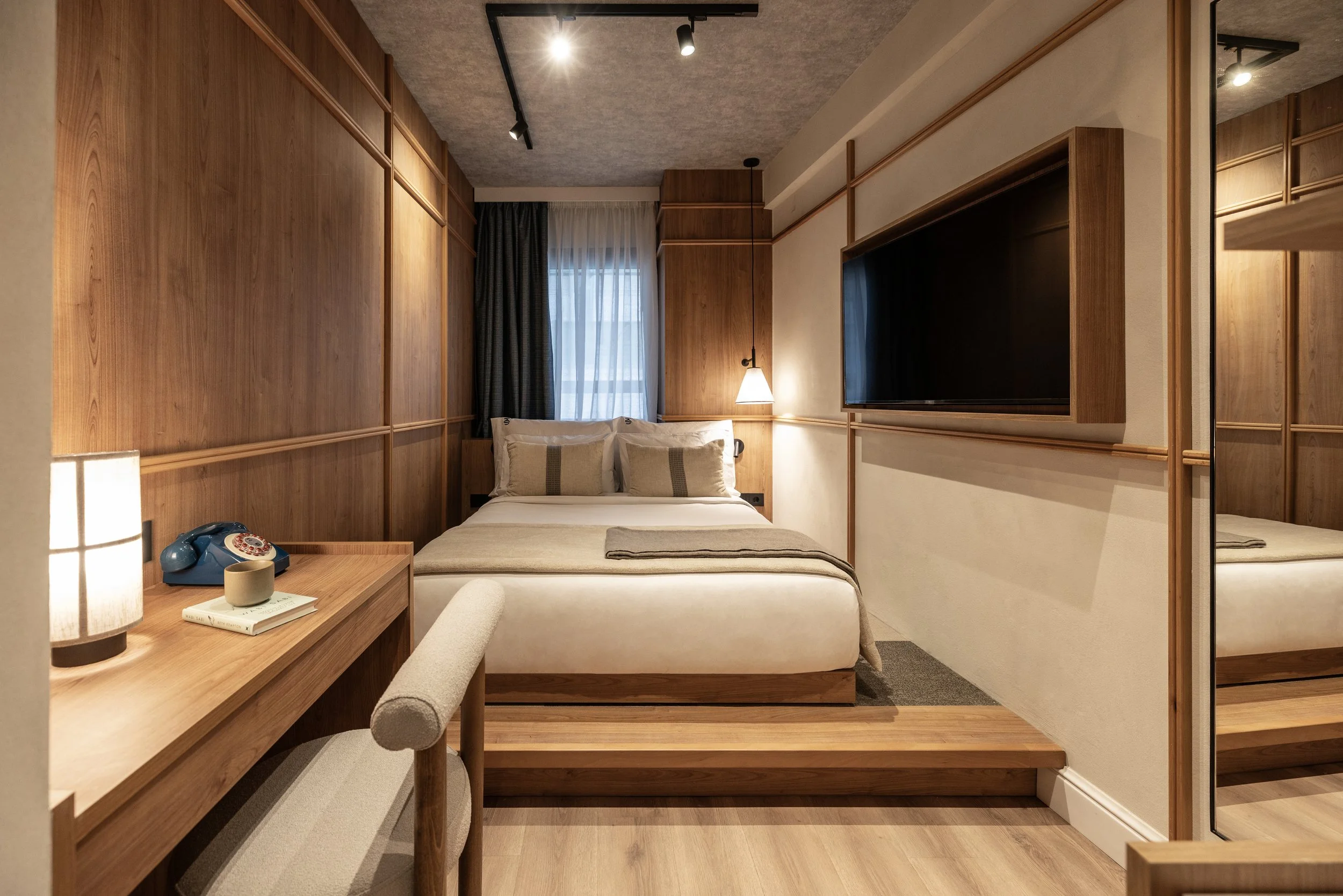 A cozy hotel room with a double bed, wooden wall paneling, a window with curtains, a desk with a blue rotary phone, a lamp, and a wall-mounted television.
