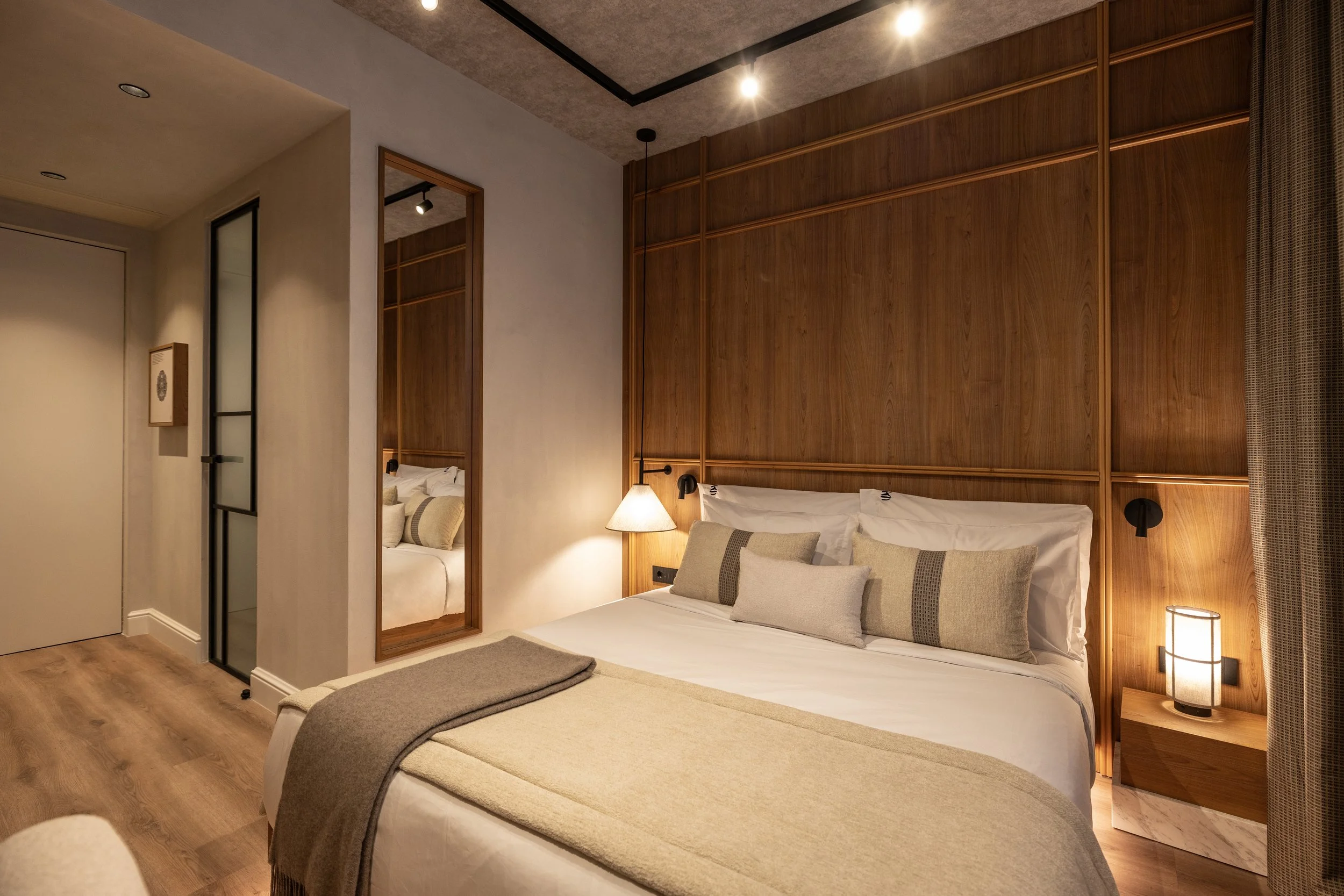 A cozy bedroom with wooden wall paneling, a bed with white sheets and beige pillows, a bedside table with a beige lamp, and a mirror on the right side.