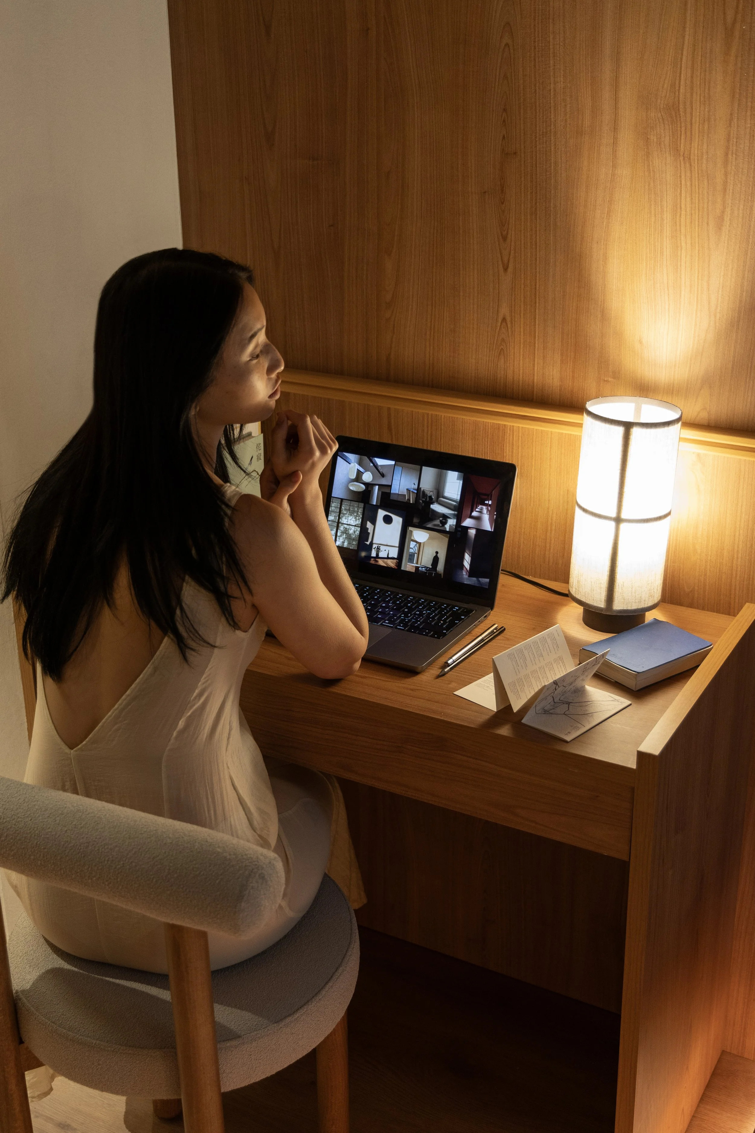 A woman with long dark hair sitting at a wooden desk in a dimly lit room, looking at a laptop screen displaying interior decor images, with a table lamp, books, and informational cards on the desk.