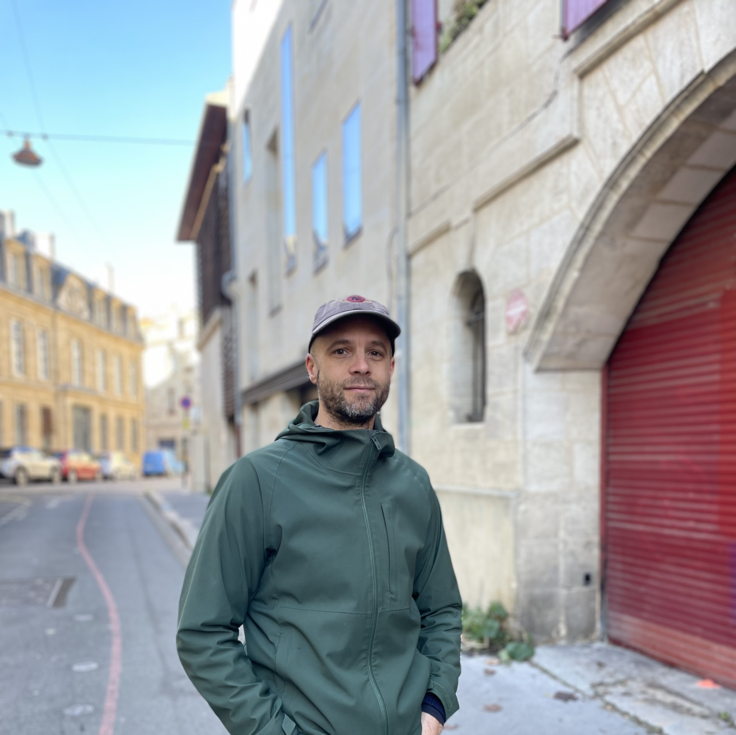 Un homme portant une veste verte et une casquette se tient dans une rue urbaine au bord d'un trottoir, avec des bâtiments anciens en pierre et un ciel clair en arrière-plan.