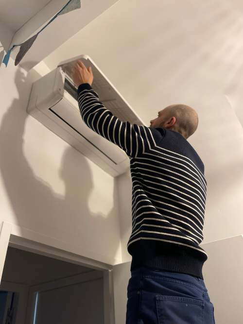 Un homme en pull à rayures ajuste un appareil de climatisation murale dans une pièce.