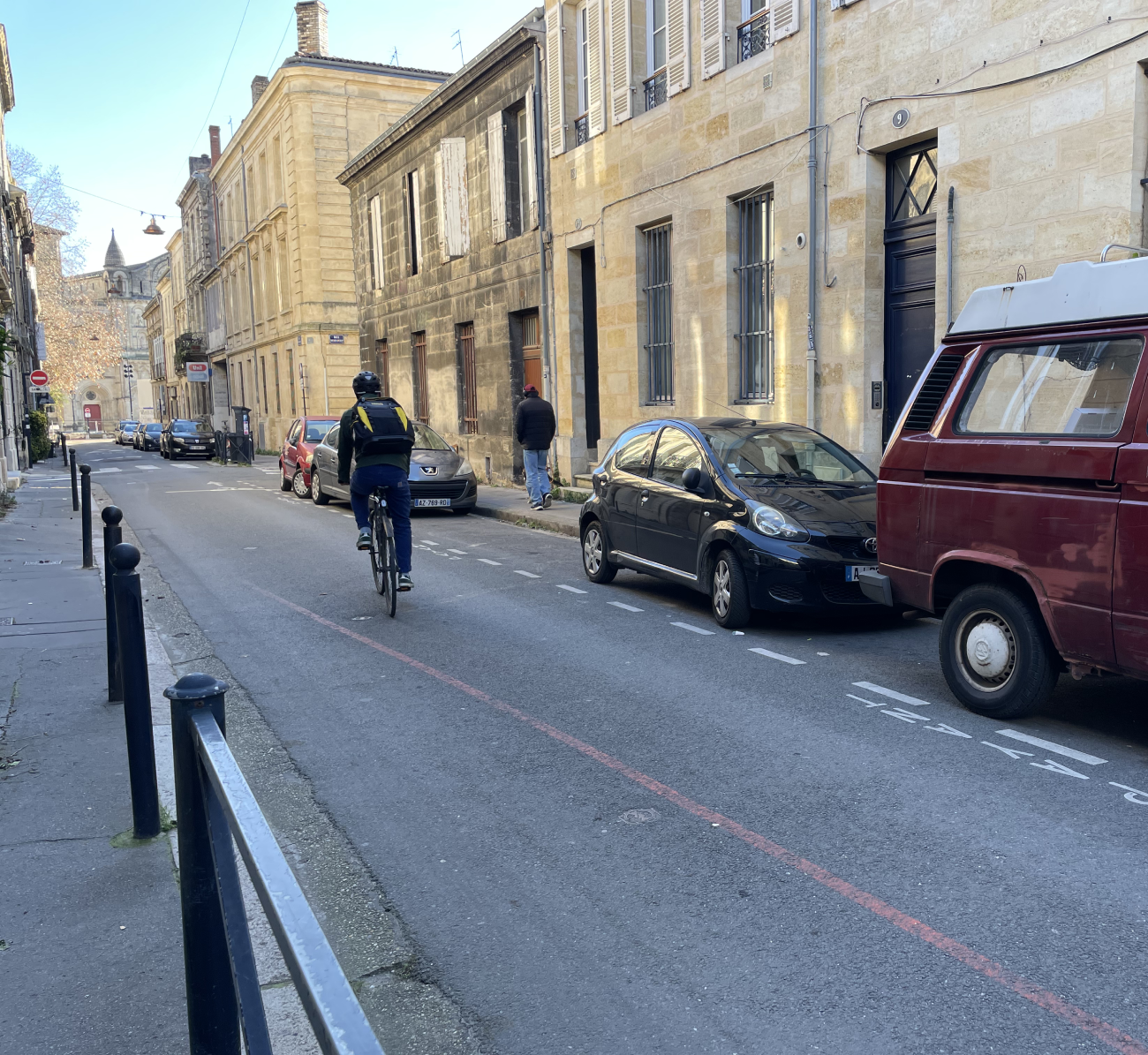 Une rue pavée de Bordeaux avec des bâtiments en pierre, des voitures stationnées le long du trottoir et un cycliste portant un casque et un sac à dos qui circule sur la voie et qui se rend sur un chantier en dépannage.