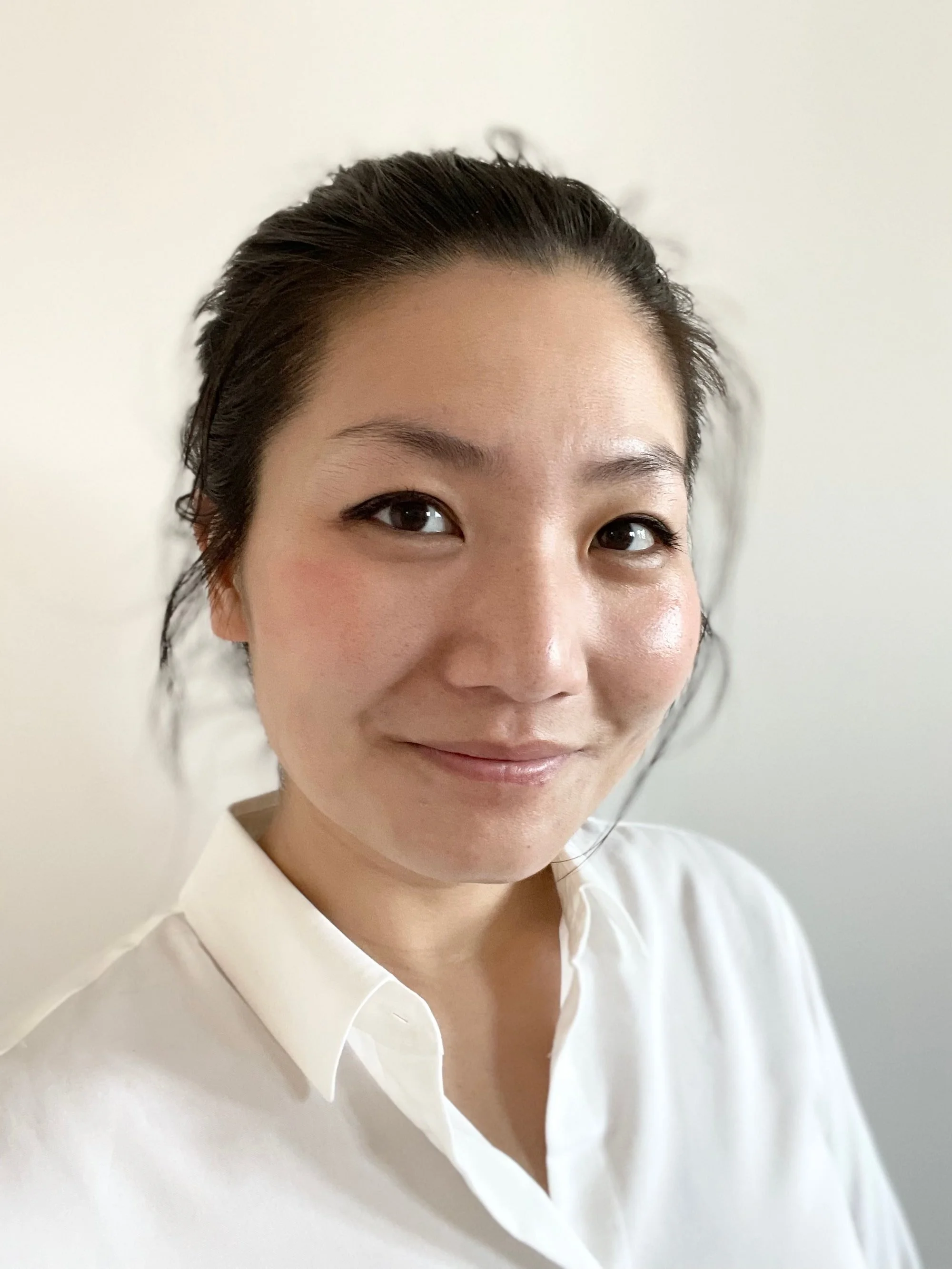 Close-up photo of a smiling Asian woman with short dark hair, wearing a white collared shirt, standing against a plain light-colored background.