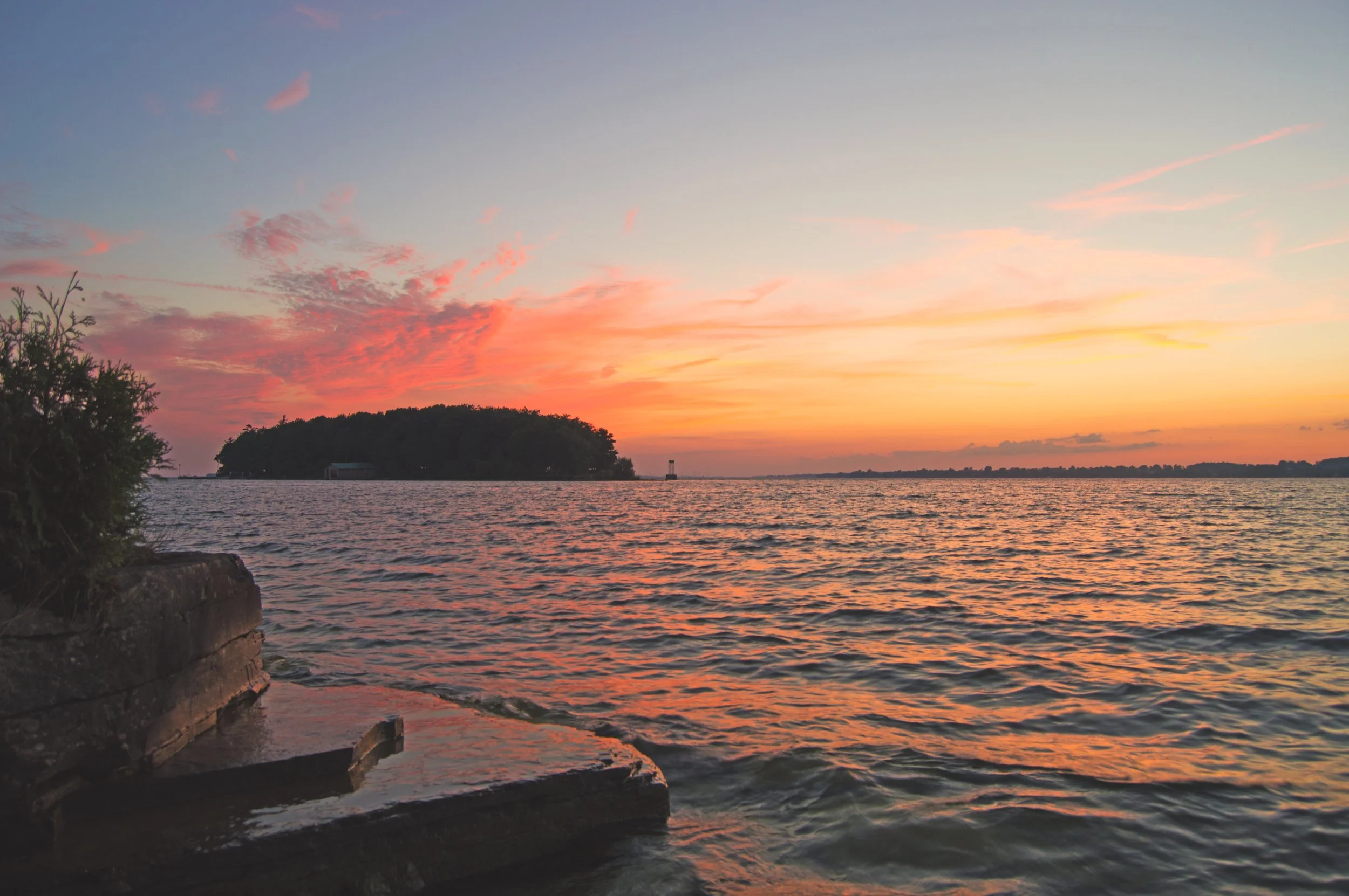 Sunset over a body of water with colorful clouds in the sky, a small island in the distance, and a rocky shoreline in the foreground.