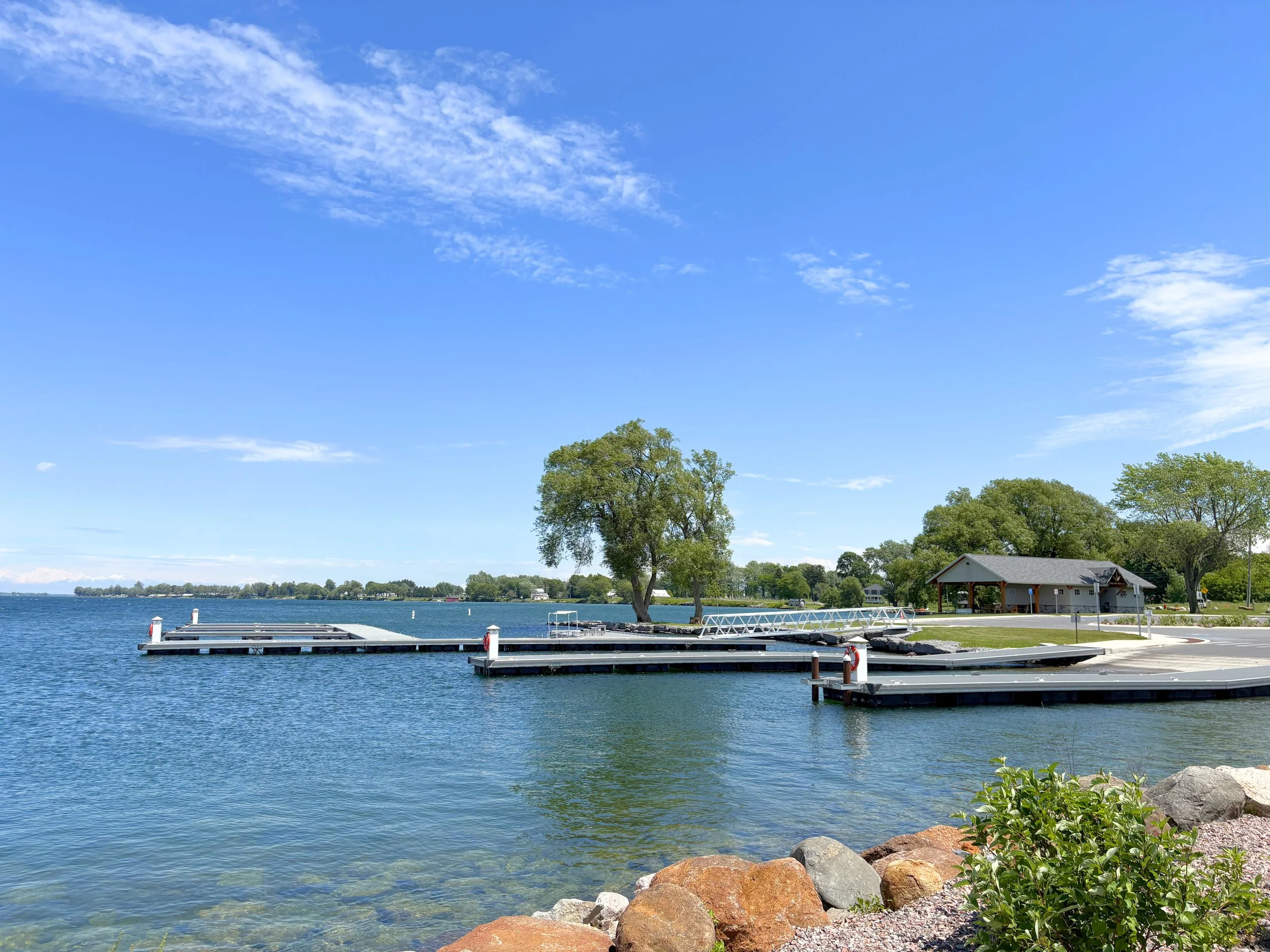 A lakeside view with a clear blue sky, scattered clouds, a dock extending into the water, trees along the shoreline, and a small building near the trees.