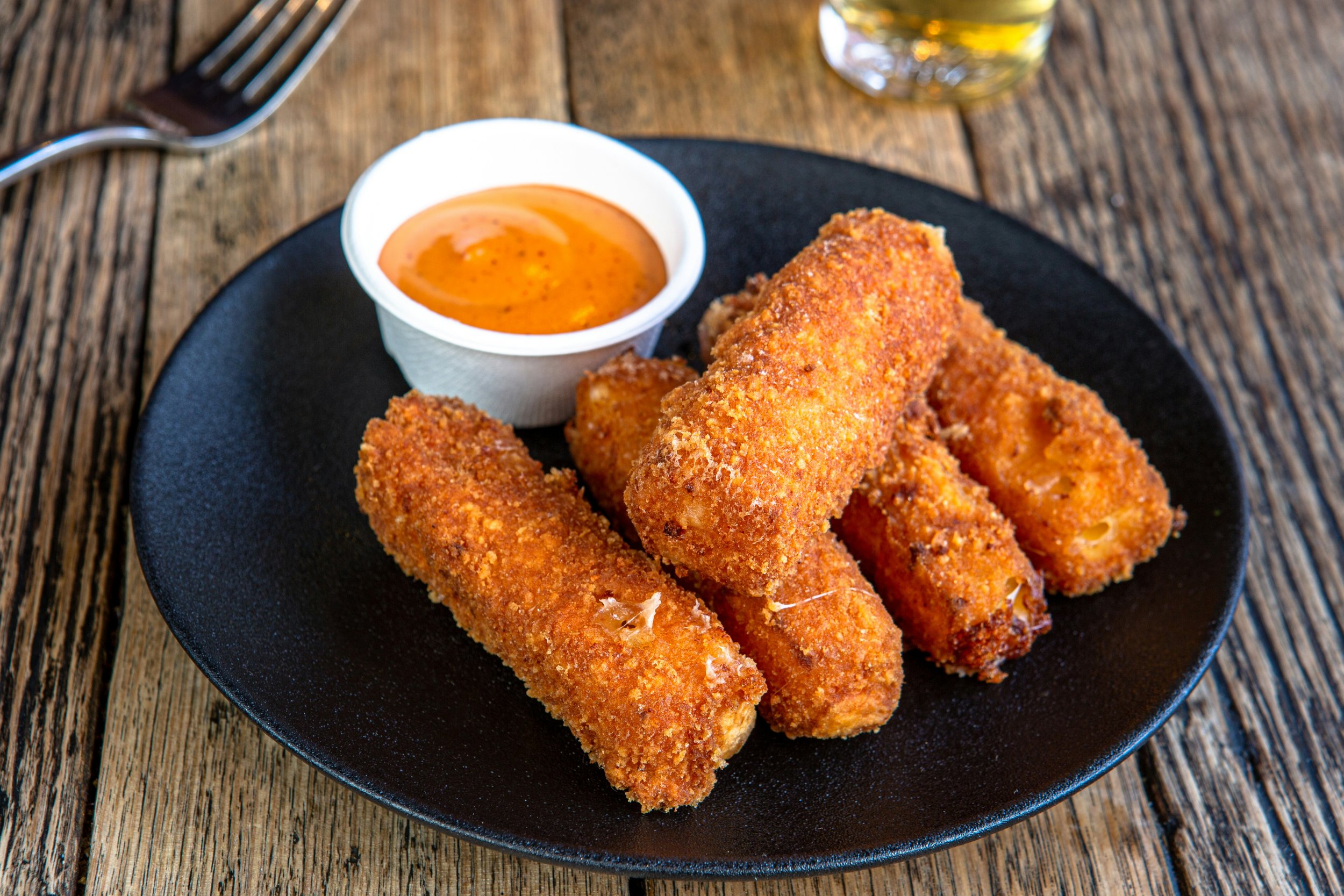 A black plate with four fried mozzarella sticks, a small bowl of marinara sauce, and a glass of beer on a wooden table.
