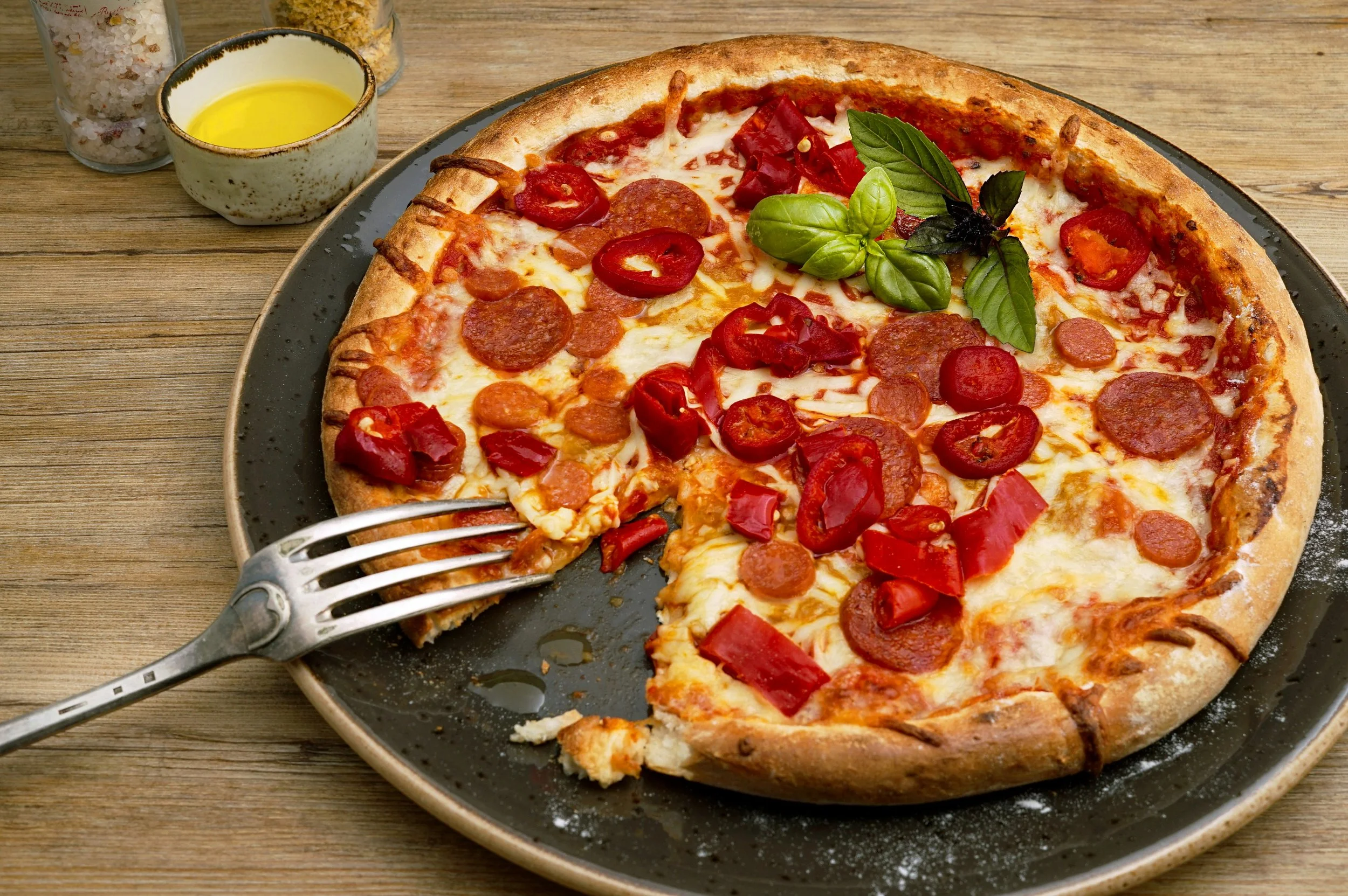A pepperoni and red bell pepper full pizza with fresh basil and oregano on top, served on a black plate with a slice being taken out. In the background, there are bottles of oil, salt, and grated cheese.