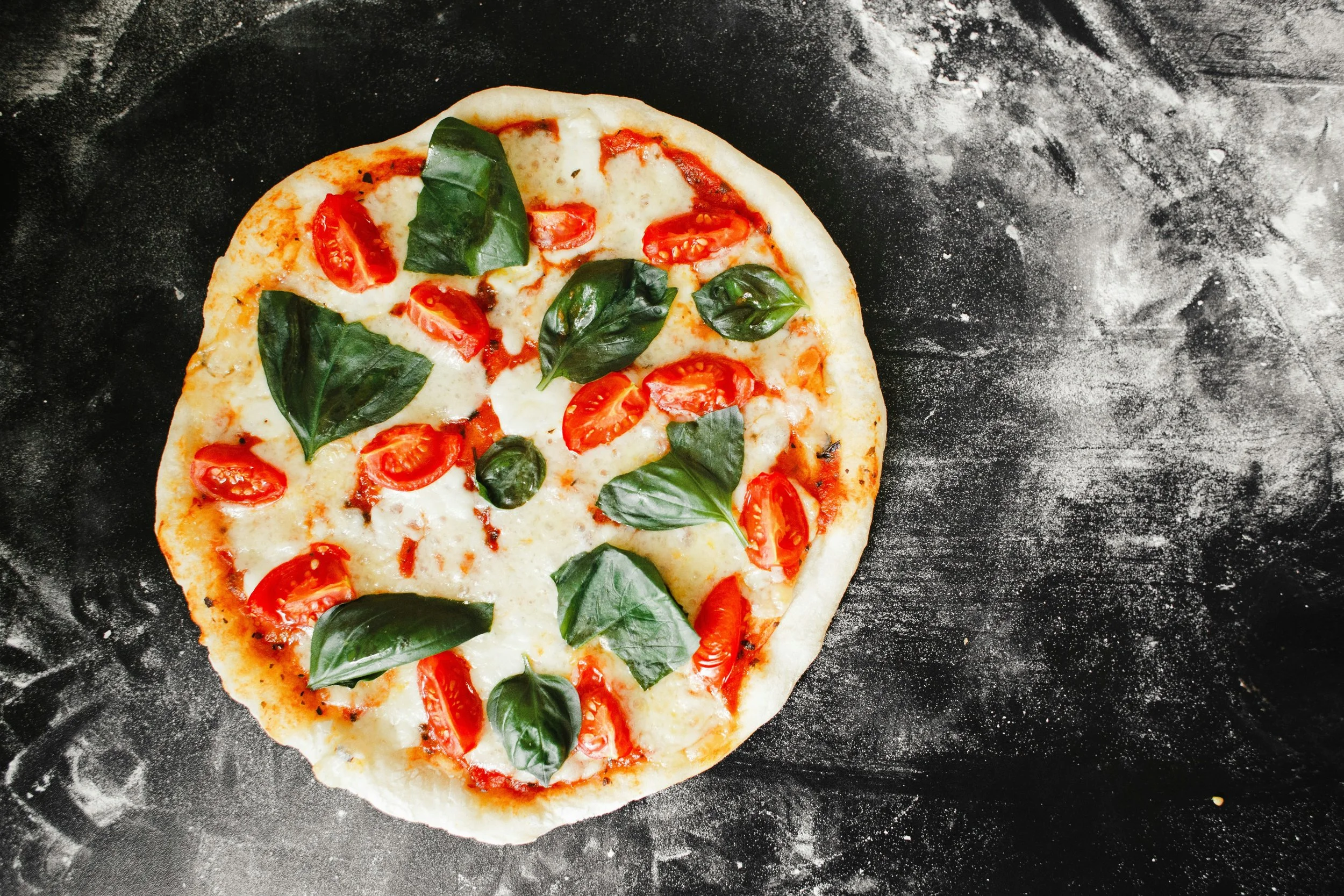 Fresh pizza with cherry tomatoes, mozzarella cheese, and basil leaves on a black surface.