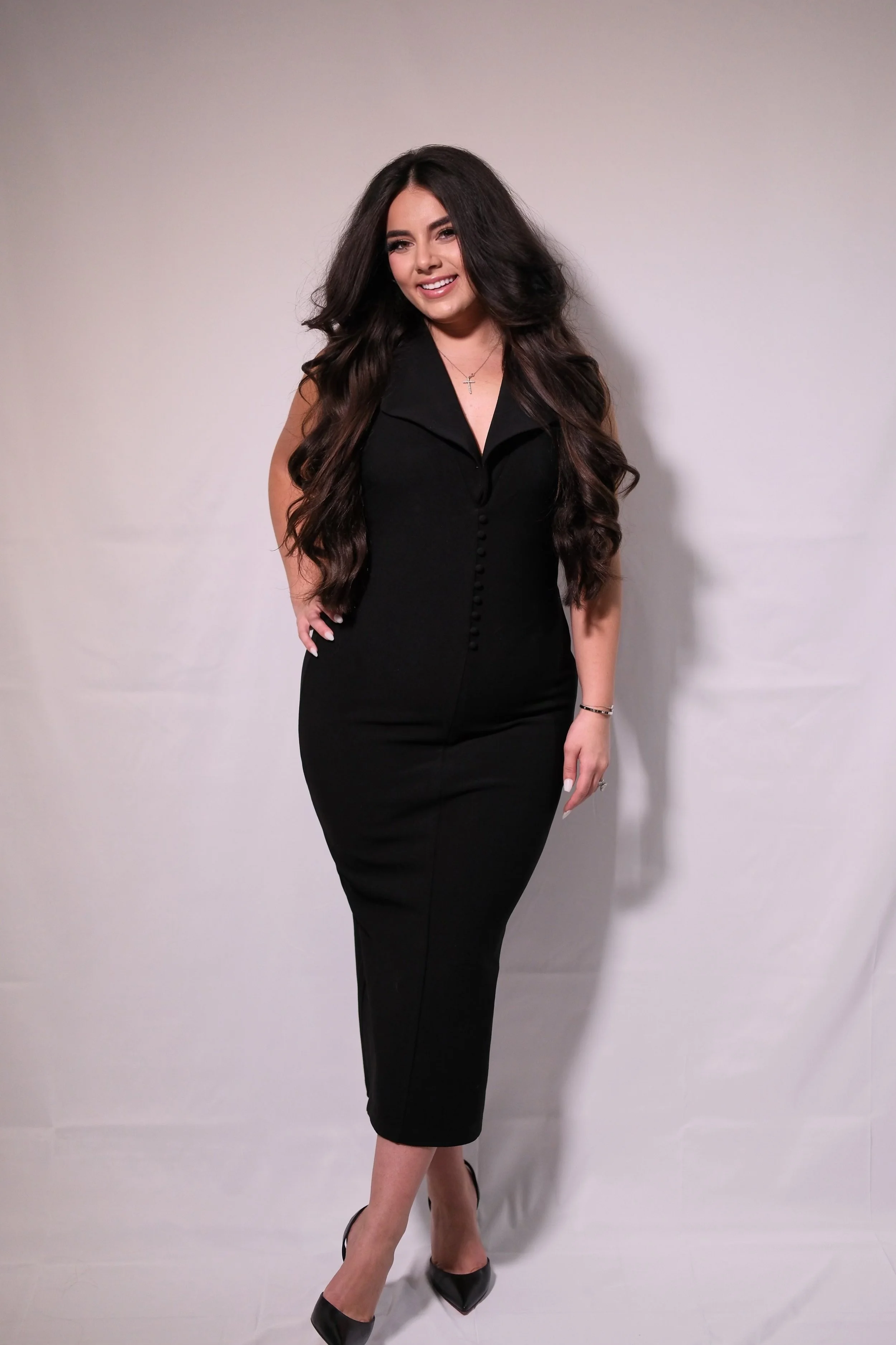 A woman with long, dark hair styled in loose waves, wearing a fitted black dress with buttons down the front, black heels, and a small necklace, standing against a plain light-colored background and smiling at the camera.