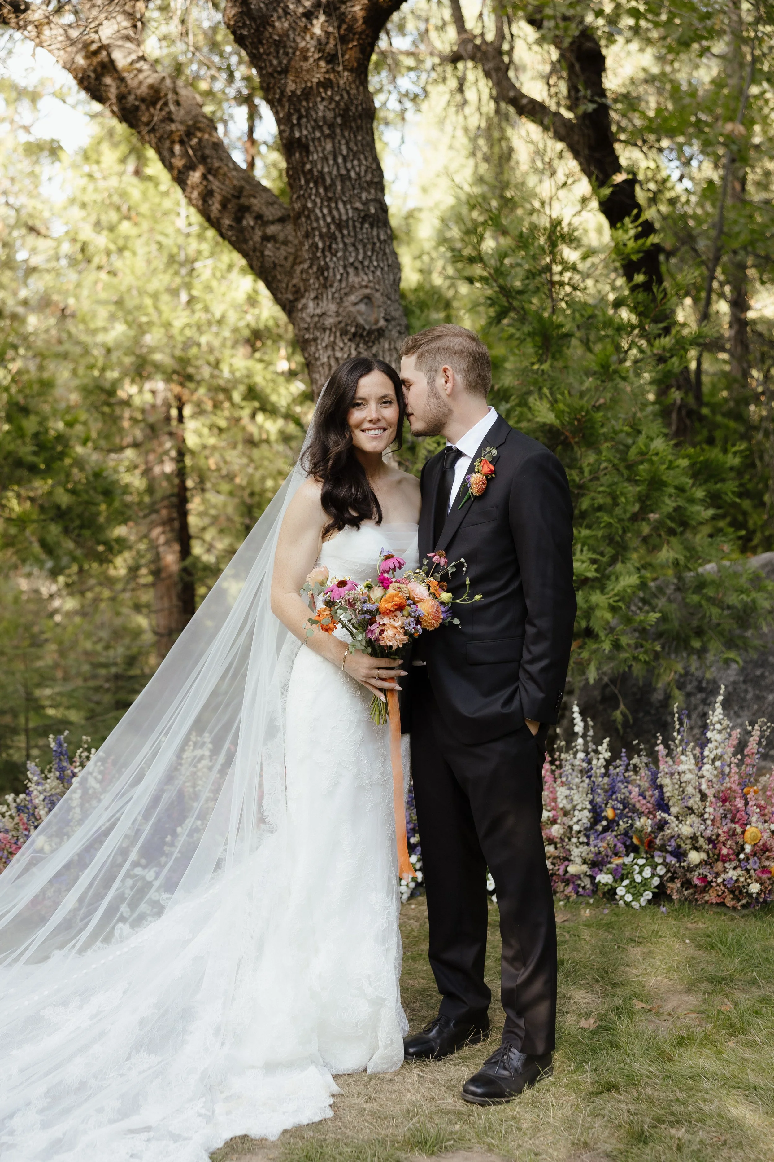 A bride and groom in wedding attire standing outdoors in a garden surrounded by trees and flowers. The bride holds a colorful bouquet, and the groom leans in to kiss her cheek.