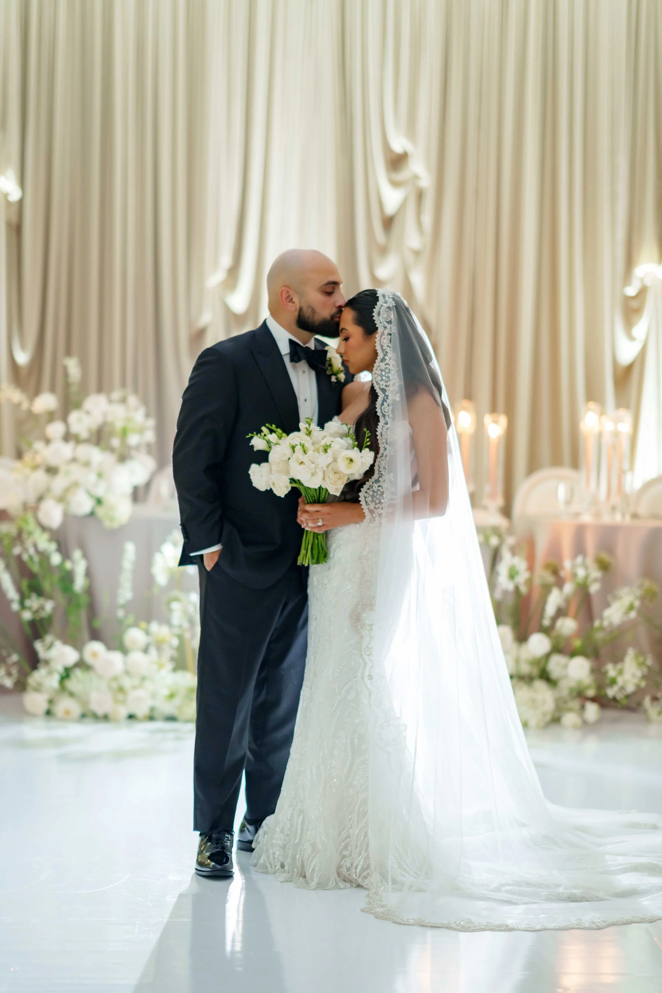 A bride and groom sharing a tender moment on their wedding day, with closed eyes and a gentle embrace, in a decorated venue with cream-colored curtains and floral arrangements.