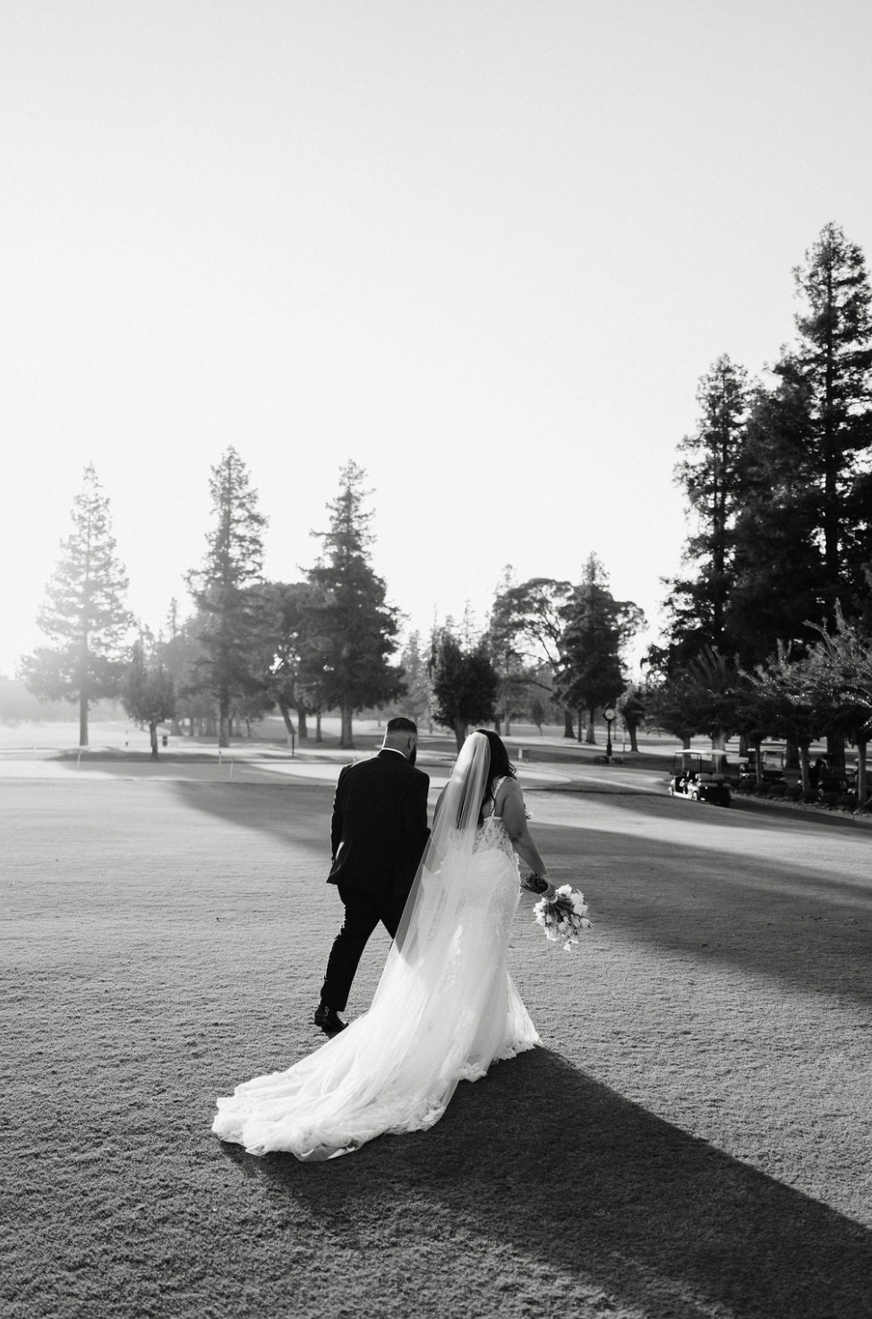 Sacramento Wedding Planner. A bride and groom walking away on a golf course, with trees and golf carts in the background, during sunset or sunrise.