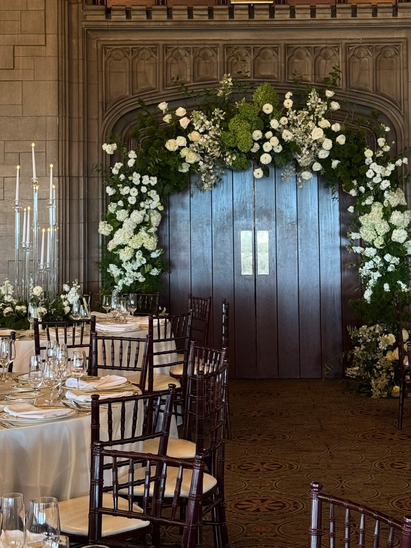 Wedding reception area with decorated arch and tables set with white tablecloths, glassware, and napkins, featuring dark wooden chairs and a large candelabrum.