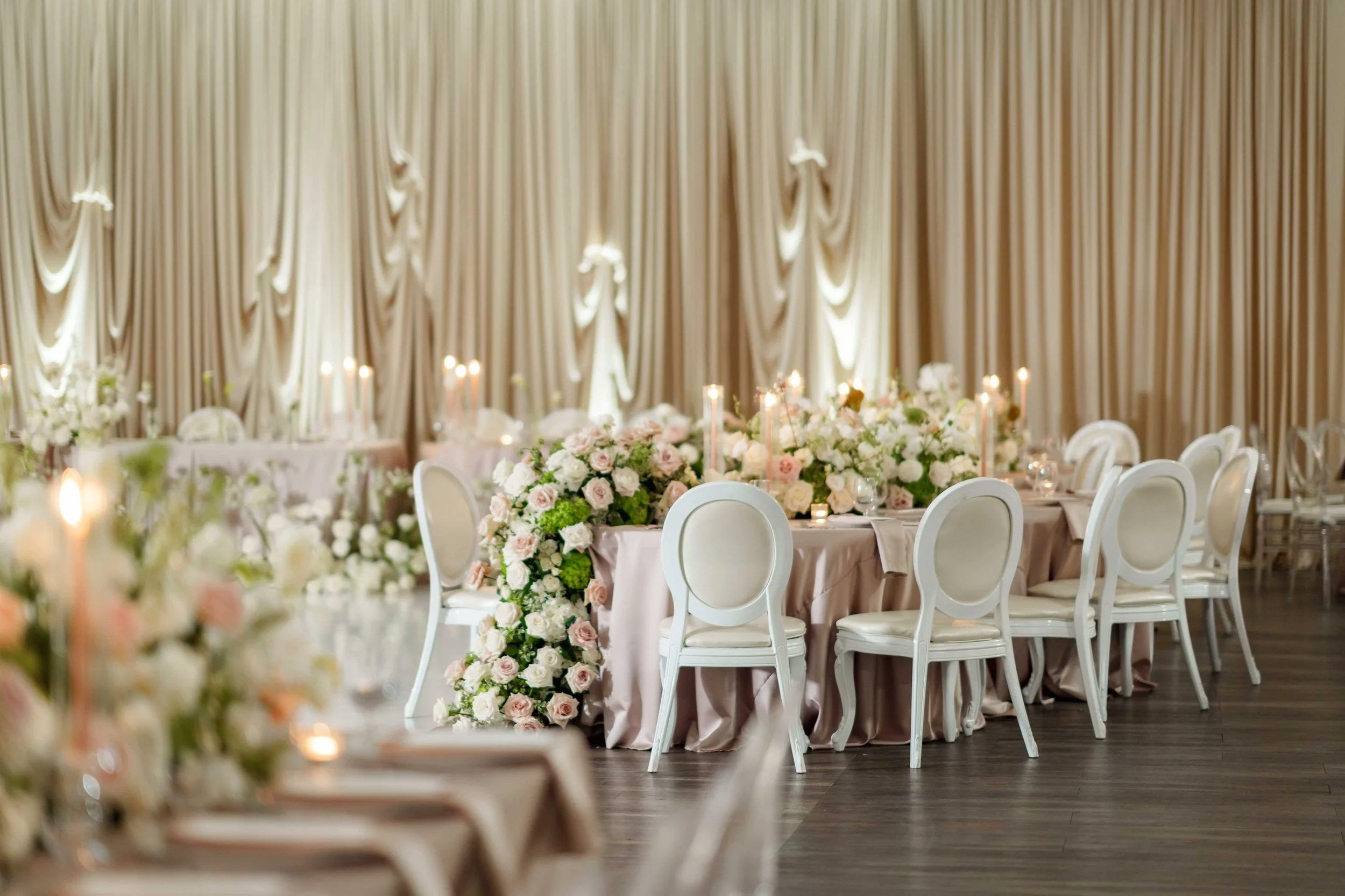 San Jose Wedding Planner. Elegant banquet hall decorated with cream and blush pink flowers and candles on round tables, with white chairs and draped curtains in the background.