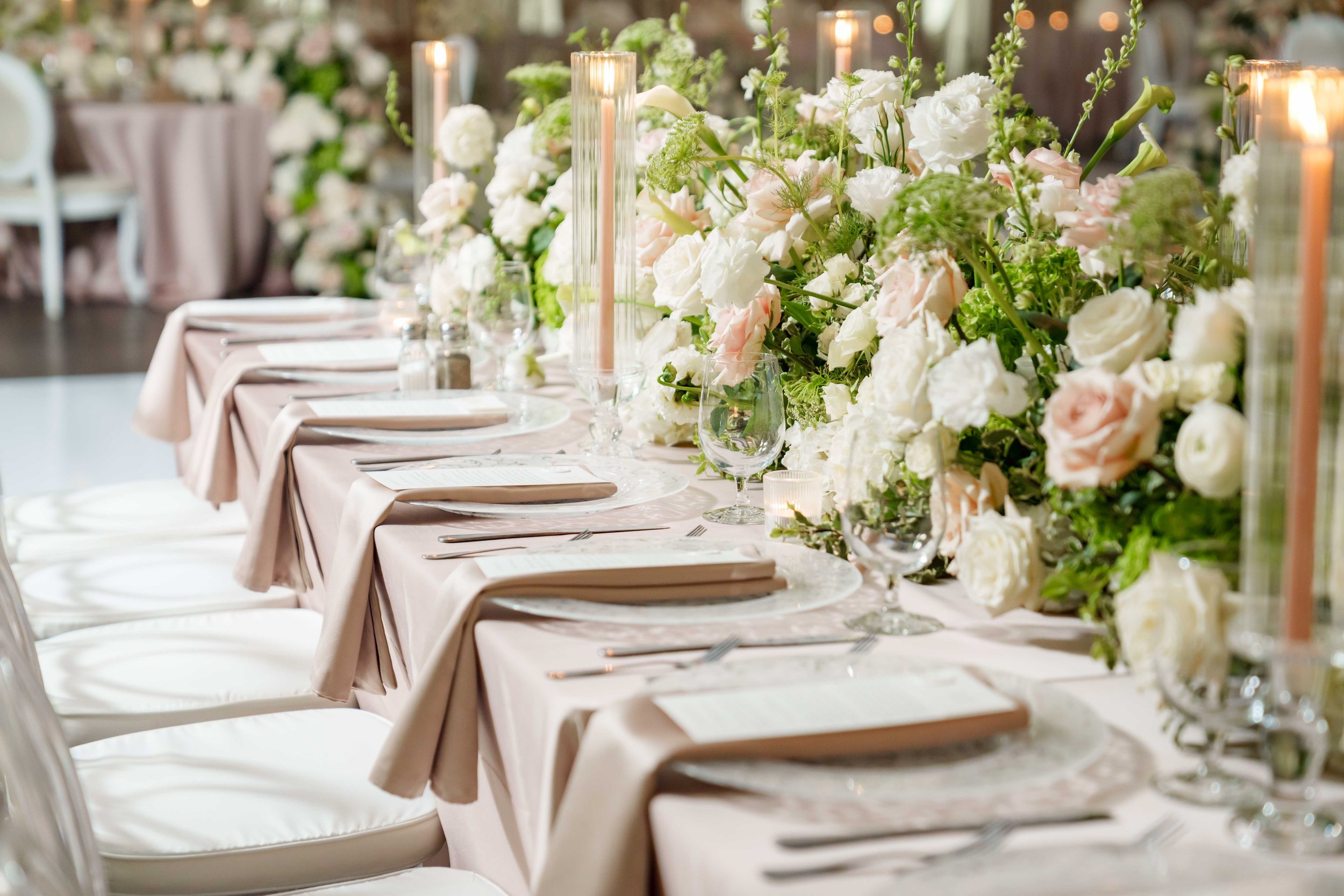 Elegant dining table decorated with a long floral centerpiece of white and blush roses, green foliage, and pink candles in glass holders, set with white plates, silverware, napkins, and glassware.