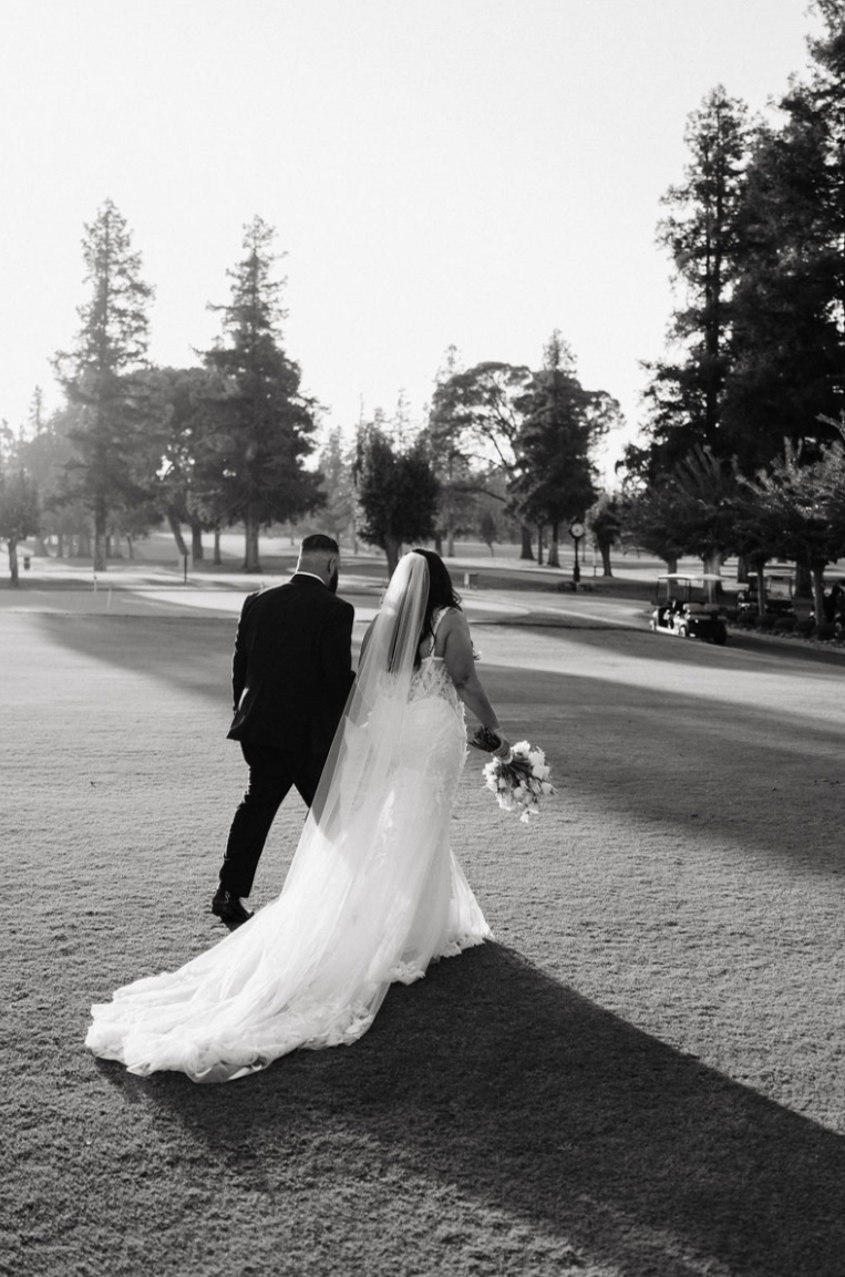A bride and groom walking together on a grassy area at sunset, the bride holding a bouquet and wearing a long train and veil, with trees and golf carts in the background.