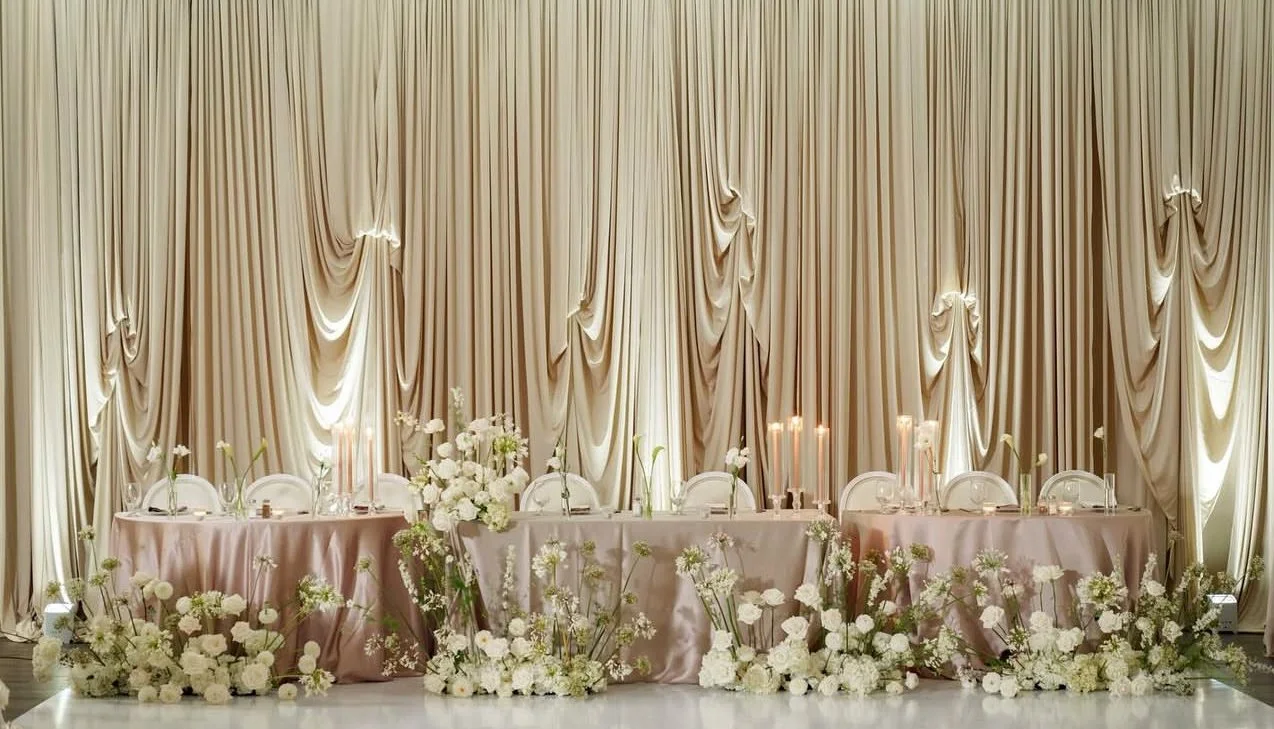 Elegant wedding table setup with a pink tablecloth, surrounded by white floral arrangements and chairs, set against a backdrop of draped beige curtains with candle centerpieces.