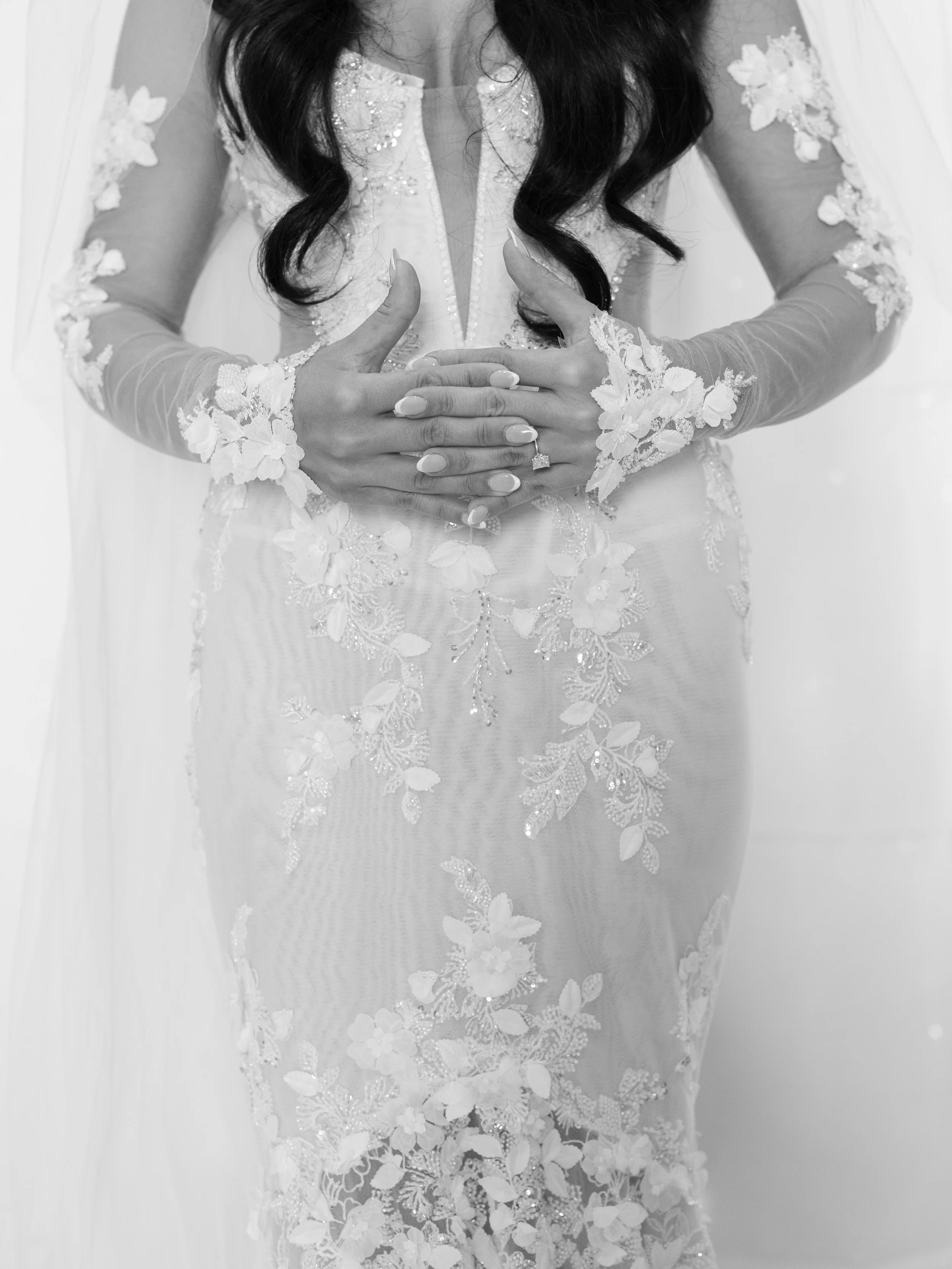 Black and white photo of a woman in an ornate, lace wedding gown with her hands over her stomach, showing a diamond ring on her finger.