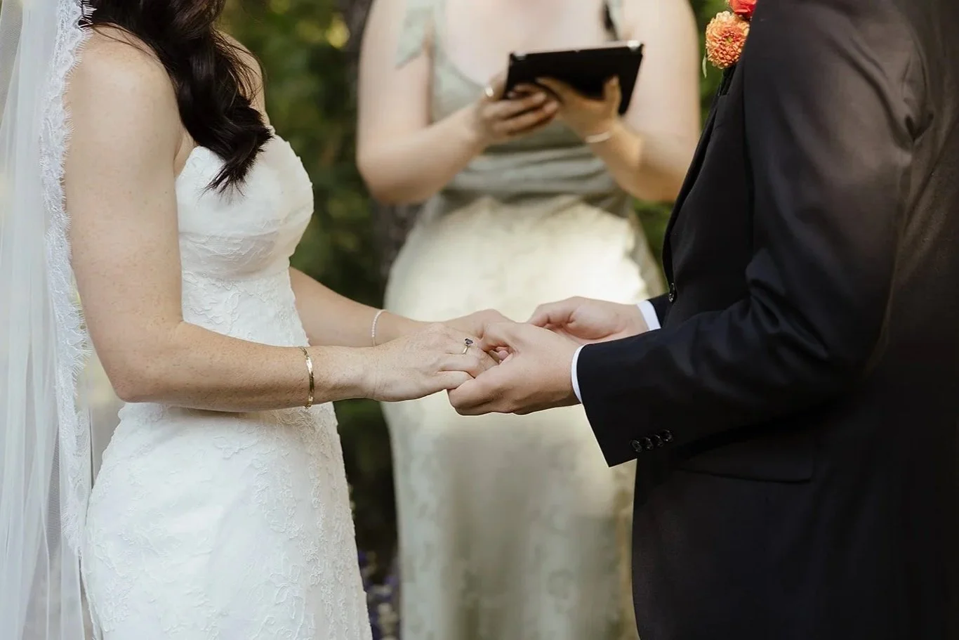 A bride and groom holding hands during a wedding ceremony outdoors, with an officiant in the background and greenery surrounding them.