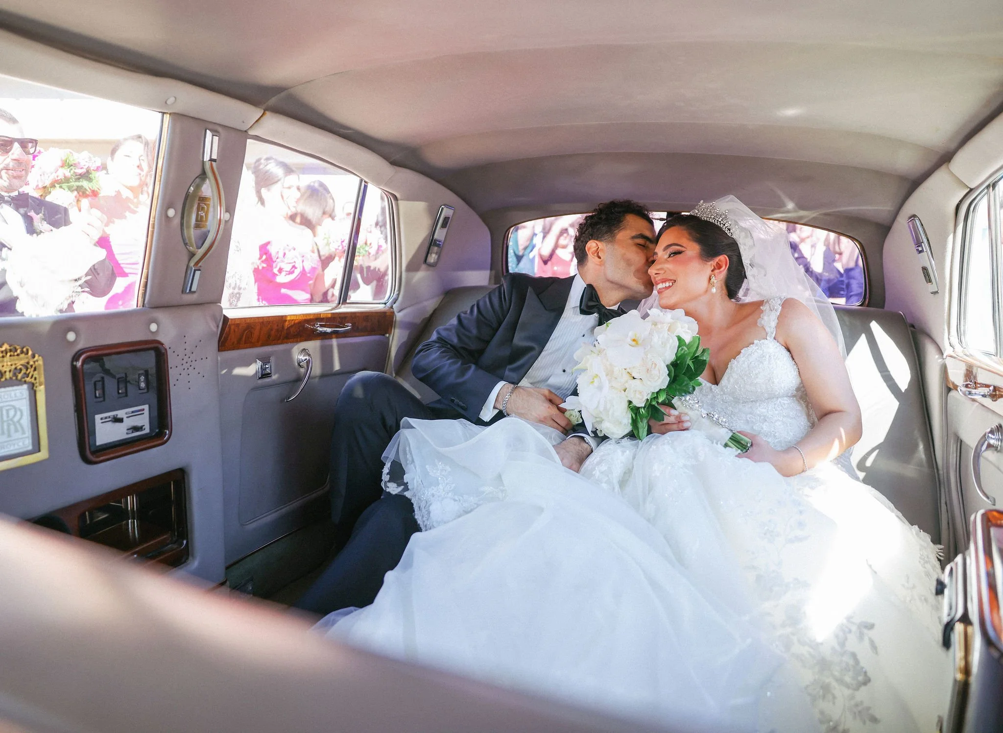 A newlywed couple sitting in the back of a vintage car, smiling and kissing while holding a bouquet of white flowers, with guests outside the car.