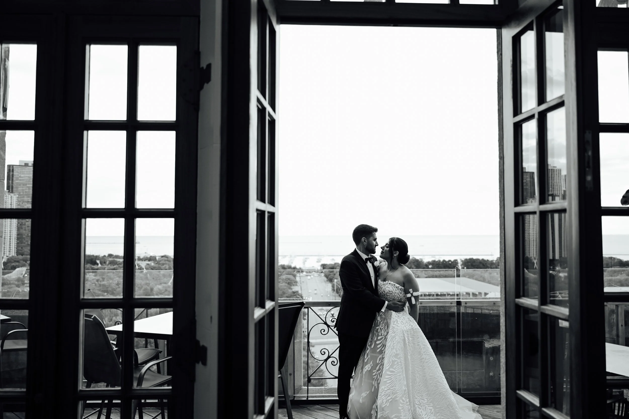 A black and white photograph of a couple in wedding attire standing on a balcony, framed by open French doors, with a cityscape and water in the background.