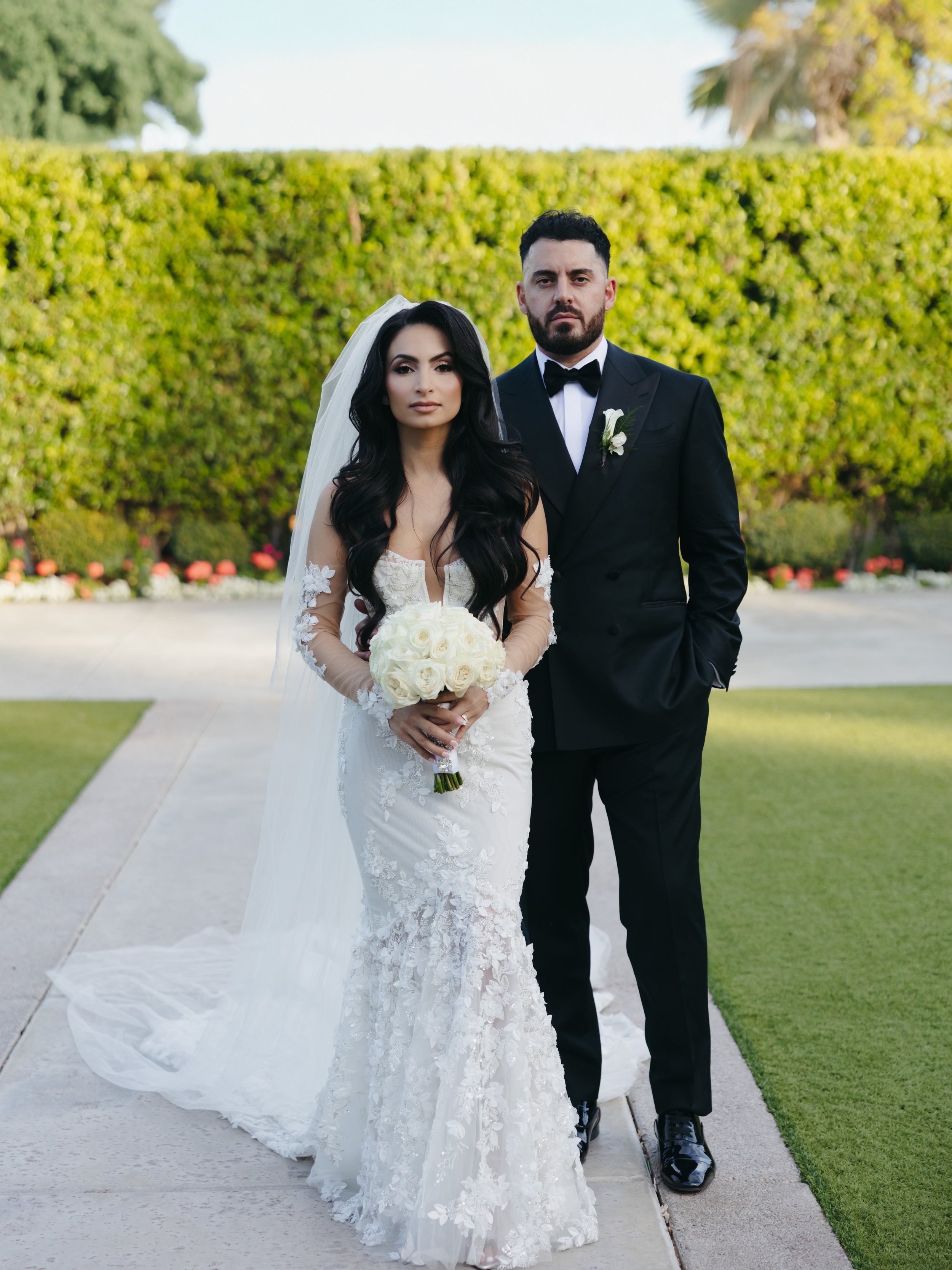 Bride and groom standing together outdoors on their wedding day, with the bride holding a bouquet of white roses.
