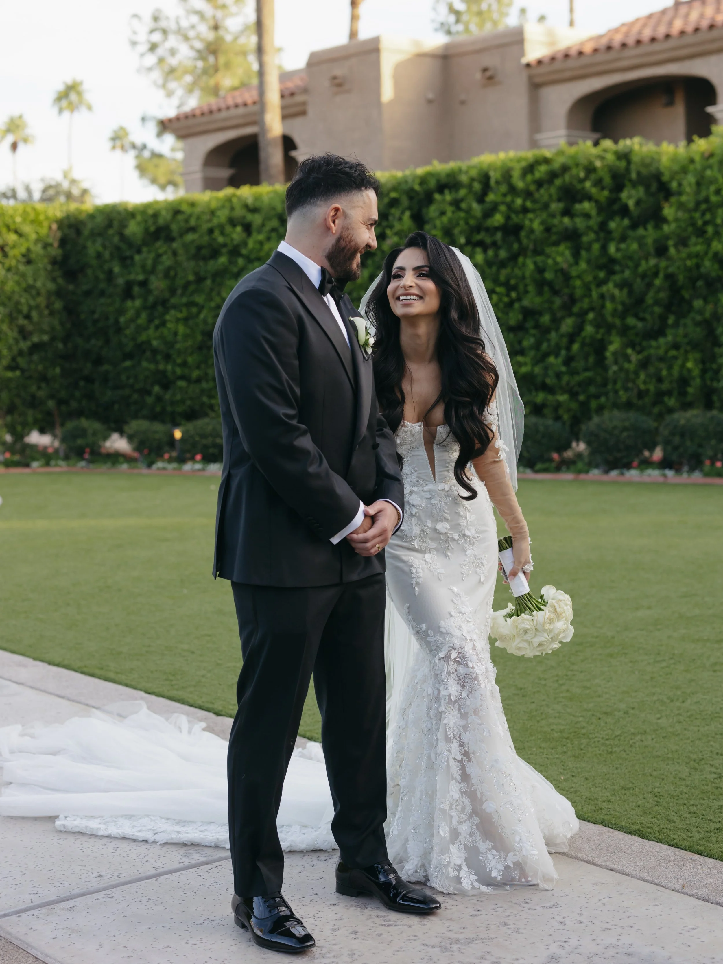 A bride and groom are smiling and looking at each other outside during their wedding. The bride is holding a bouquet of white flowers and wearing a lace wedding dress with long sleeves. The groom is dressed in a black tuxedo with a bow tie. There is greenery and a building with a red-tiled roof in the background.