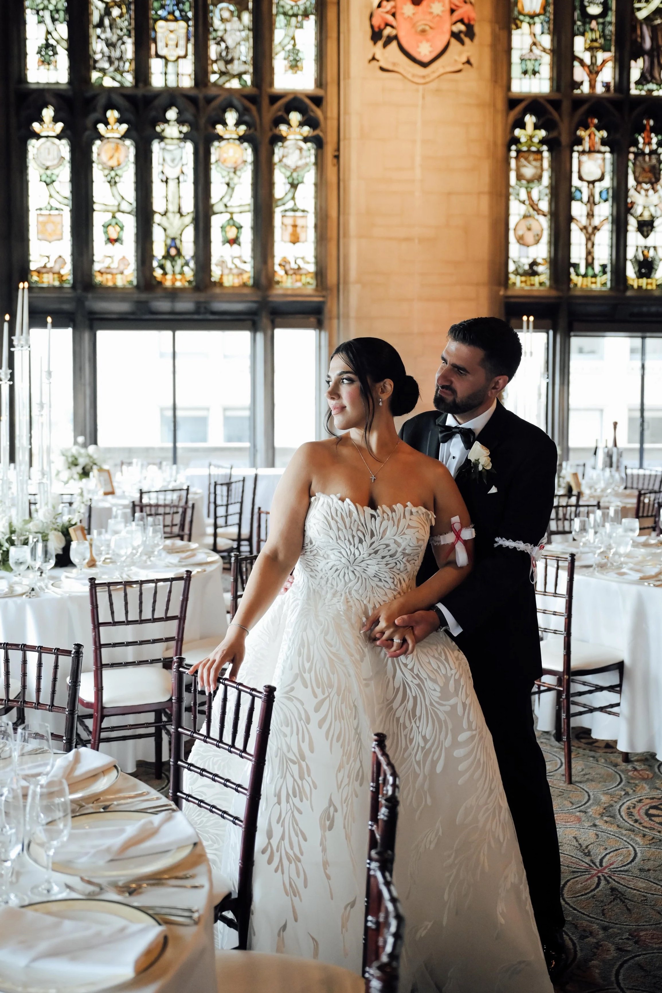 A bride and groom holding hands and dancing together in an elegant, decorated banquet hall with stained glass windows and a large crest on the wall behind them.
