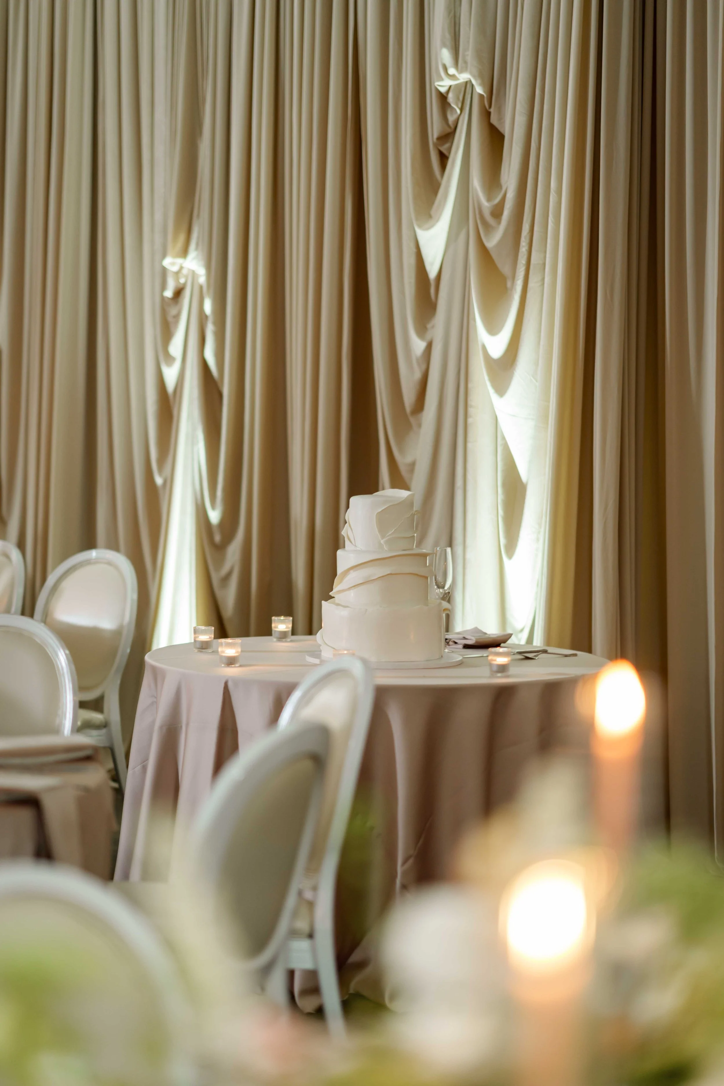 A wedding reception table with a white wedding cake surrounded by candles and elegant cream-colored curtains in the background.