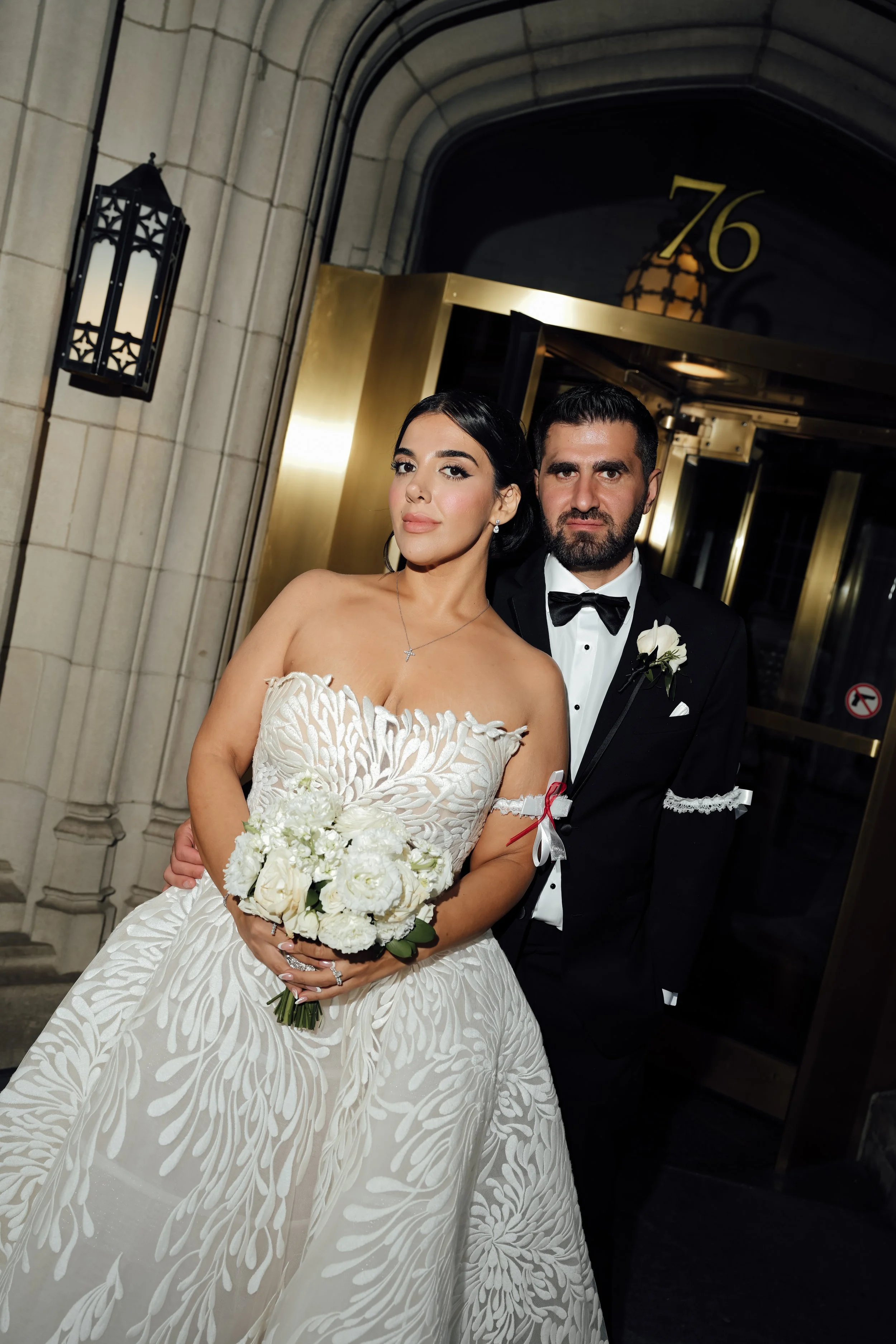 Bride in a white wedding gown holding a bouquet standing next to groom in black tuxedo with bowtie outside a building with the number 76 above the door.