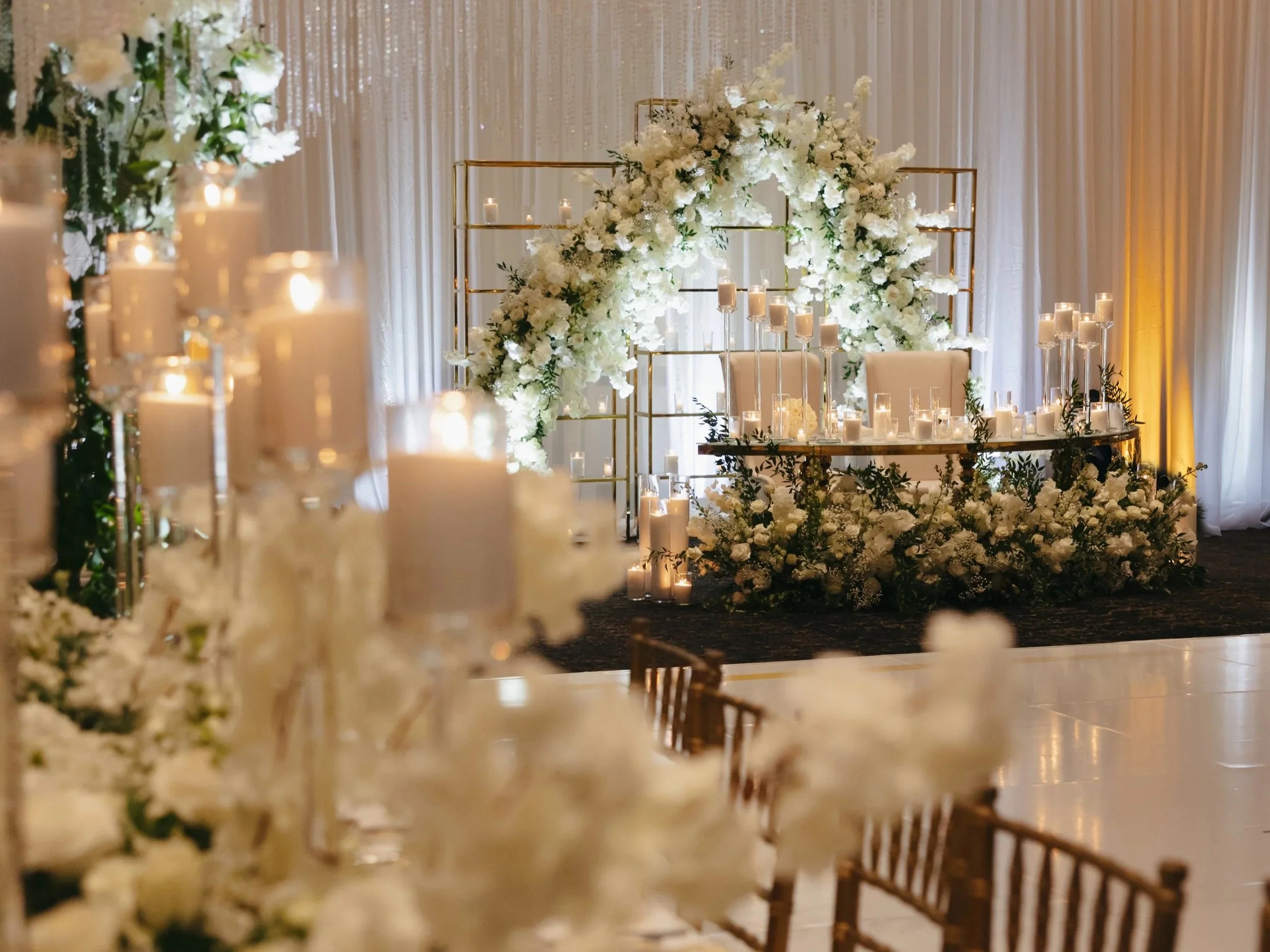 Elegant wedding reception setup with white floral arrangements and candles, featuring a floral arch and a decorated table in the background.