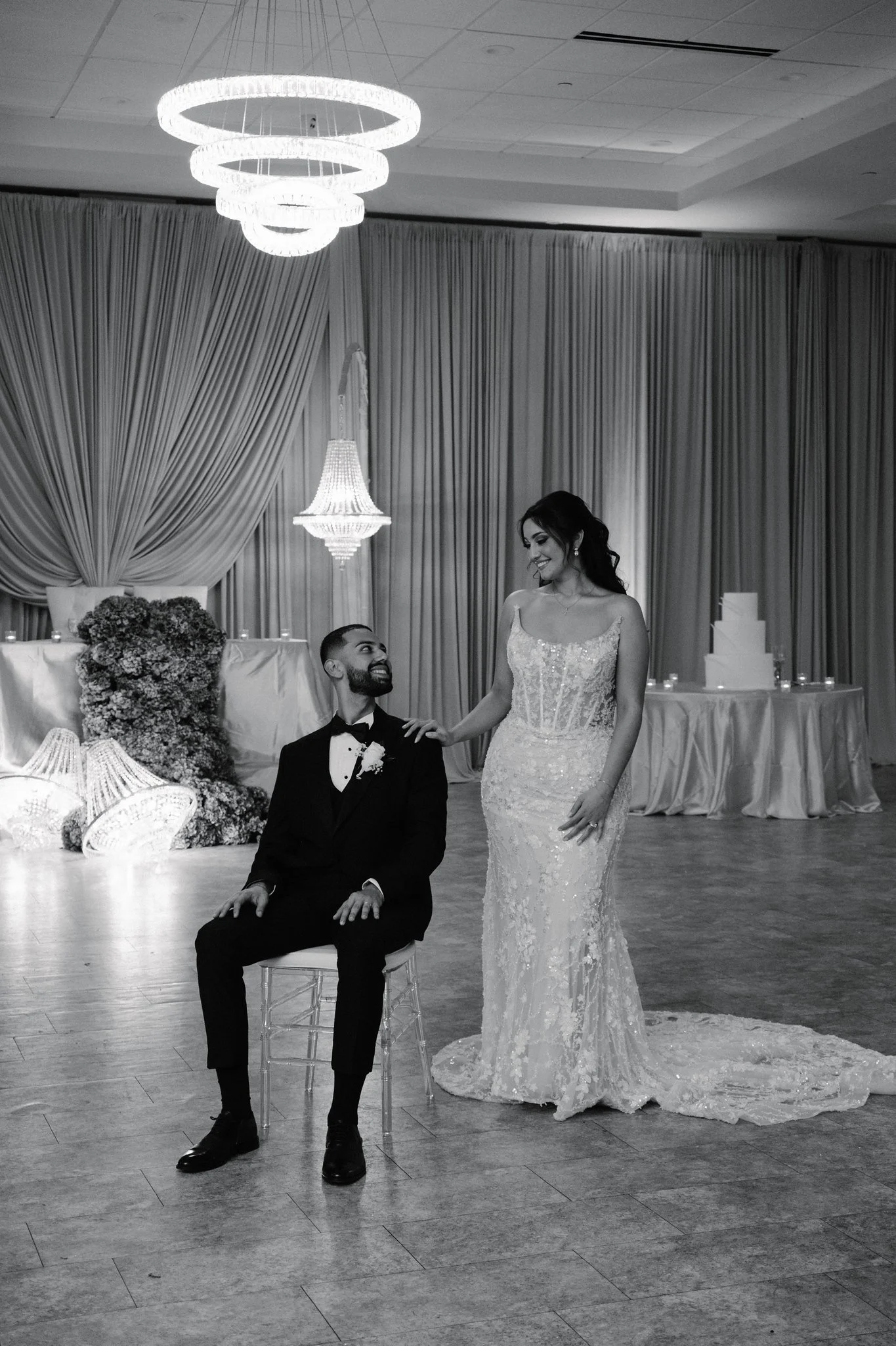 A bride and groom are in a wedding reception ballroom with curtains and a decorated backdrop; the groom is sitting on a chair, smiling at the bride, who is standing and smiling back at him.