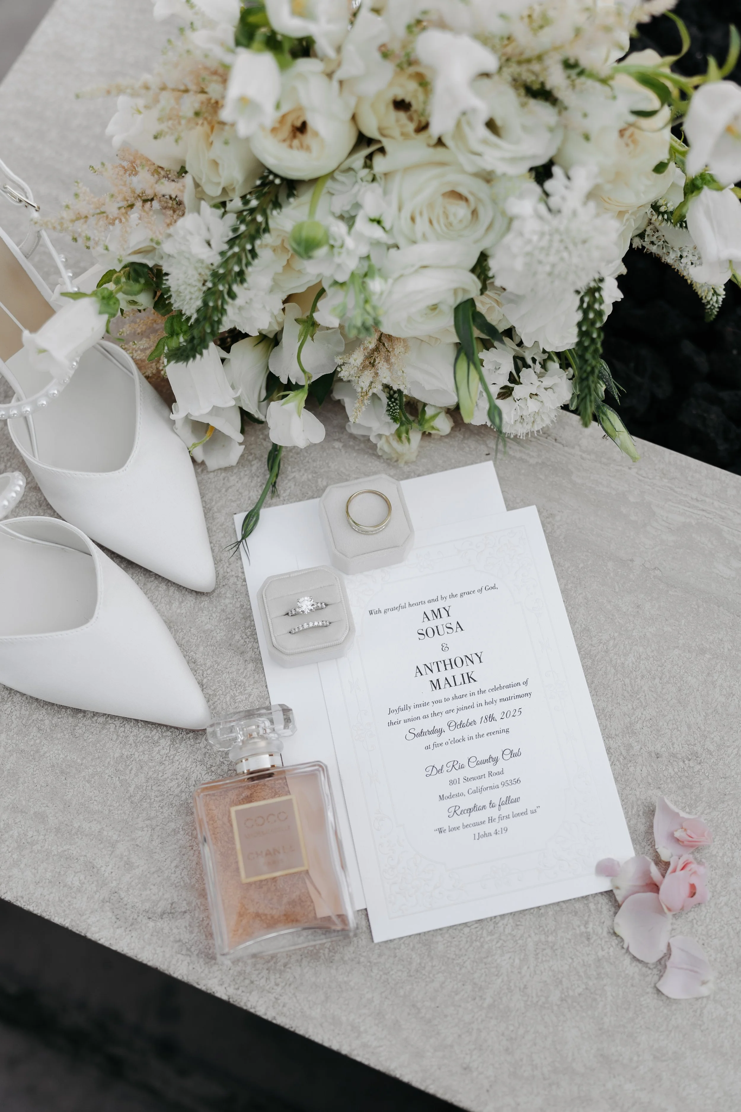 Wedding invitation with rings, white shoes, perfume, and a bouquet of white flowers on a table.