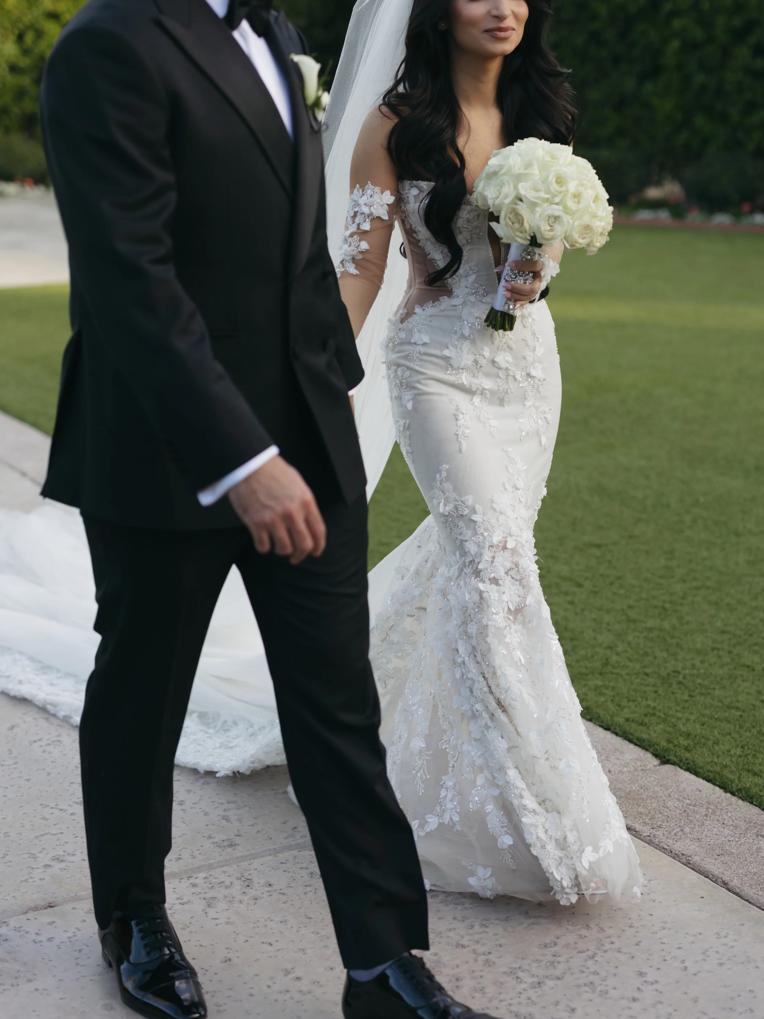 A bride in a white lace wedding dress holding a bouquet of white roses, walking beside a groom in a black tuxedo with a white boutonniere, outdoors on a paved path with green grass and trees in the background.