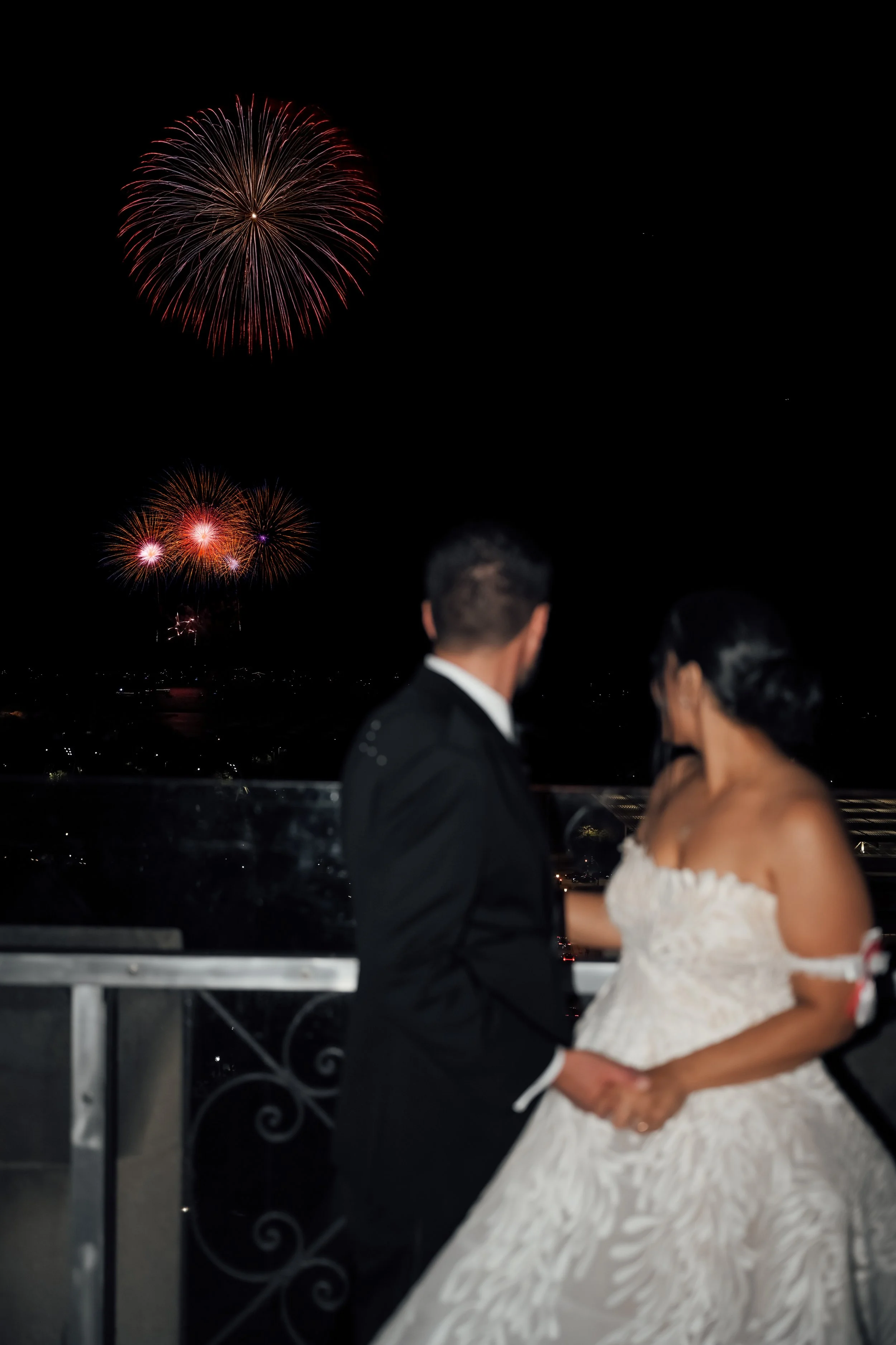 A couple dressed in wedding attire watching fireworks at night, with colorful bursts illuminating the sky.
