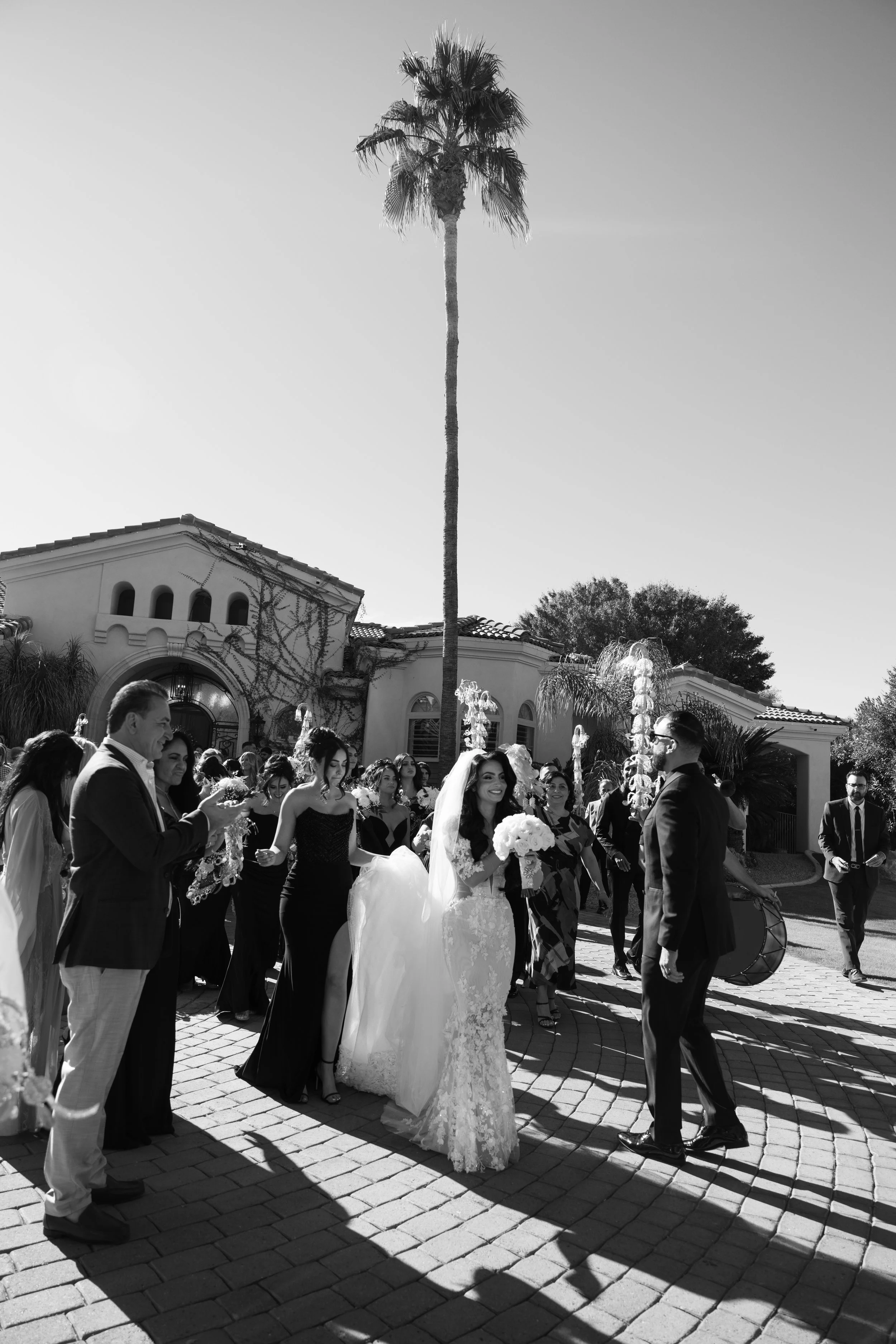 A wedding celebration outdoors with guests, a bride holding a bouquet, and a groom in a suit, under a tall palm tree and sunny sky.