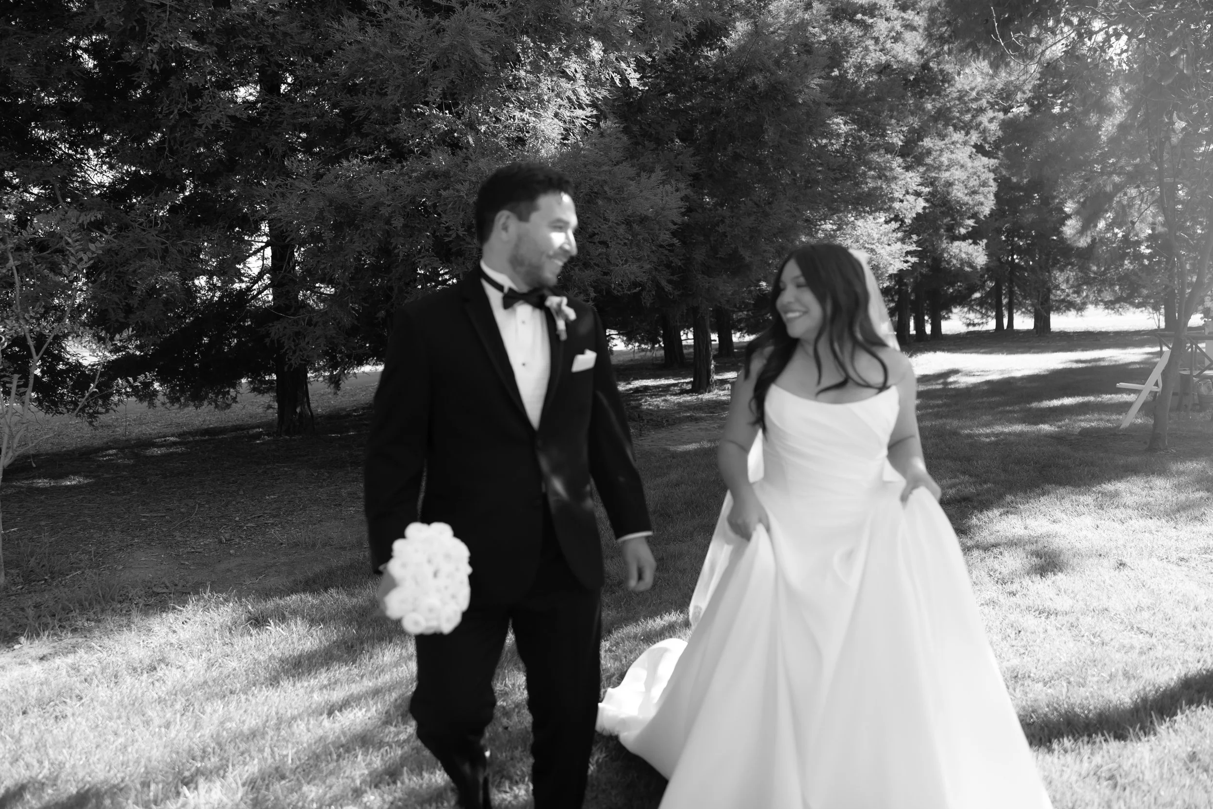 A black and white photograph of a bride in a wedding dress and a groom in a tuxedo walking outdoors on grass, smiling at each other with trees in the background.