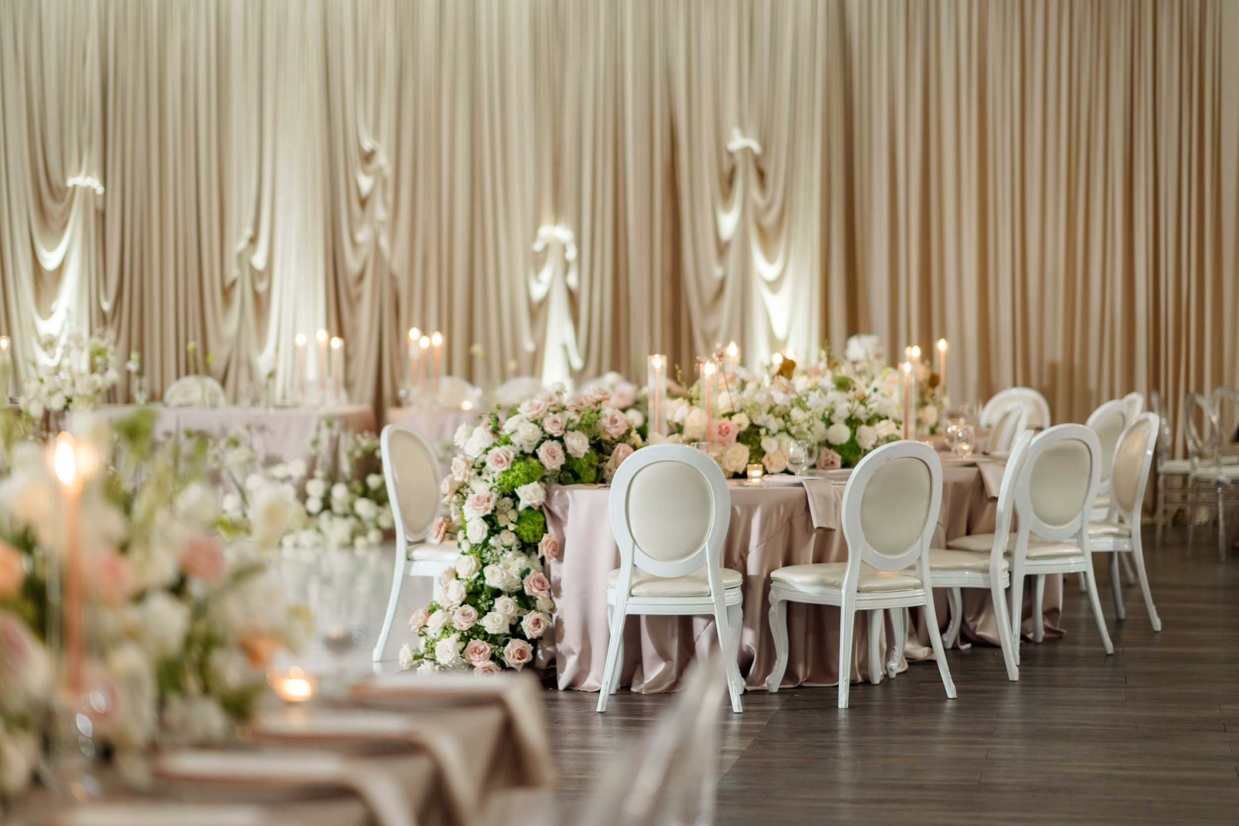 Elegant banquet hall decorated with floral arrangements and candlelit centerpieces, cream-colored drapes, and white chairs around a pink tablecloth.