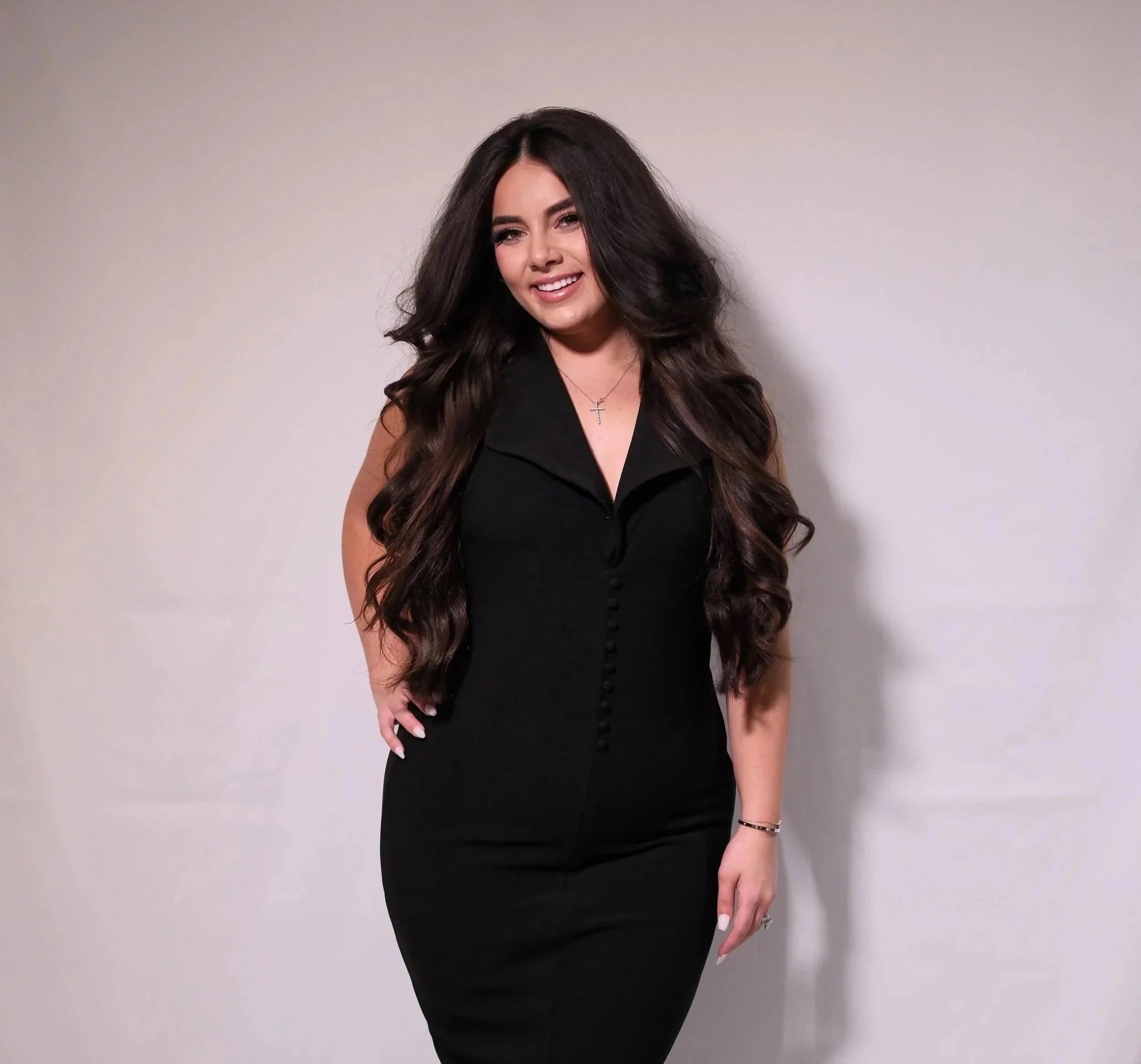 A young woman with long, dark brown hair and a bright smile, wearing a sleeveless black dress with buttons down the front, standing against a plain off-white wall.
