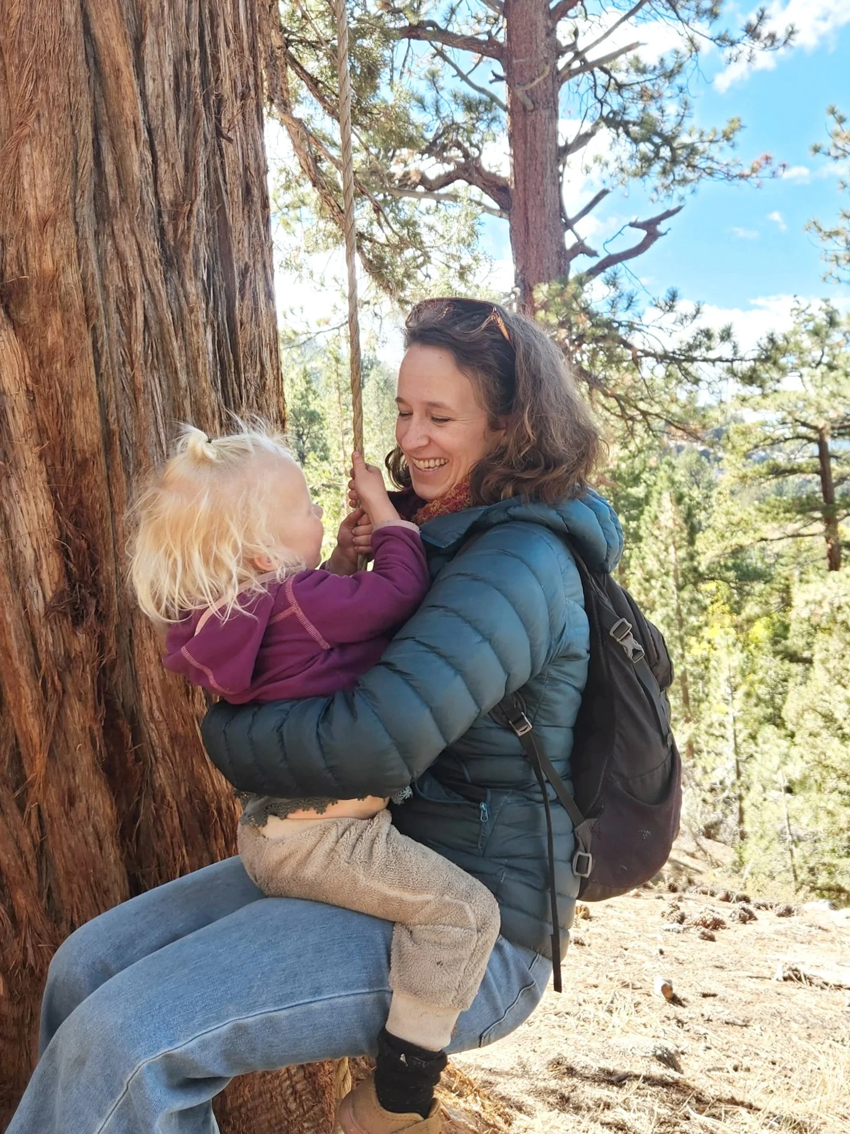 A woman and a young girl are smiling and playing on a tree swing in a forest with tall trees and blue sky.