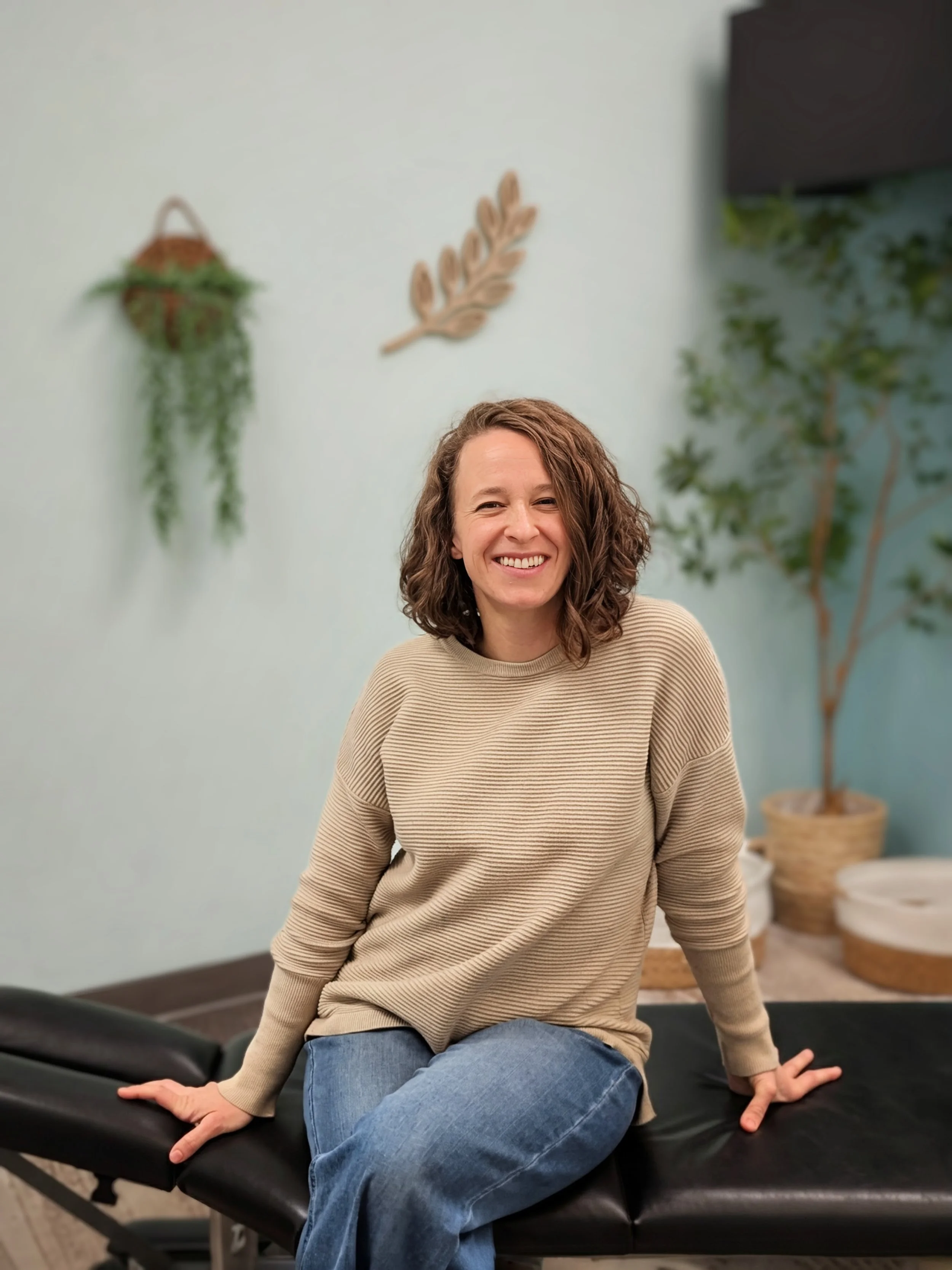 A woman with curly brown hair, wearing a beige sweater and jeans, smiling while sitting on a black massage table in a room with light blue walls, decorated with wall art and indoor plants.
