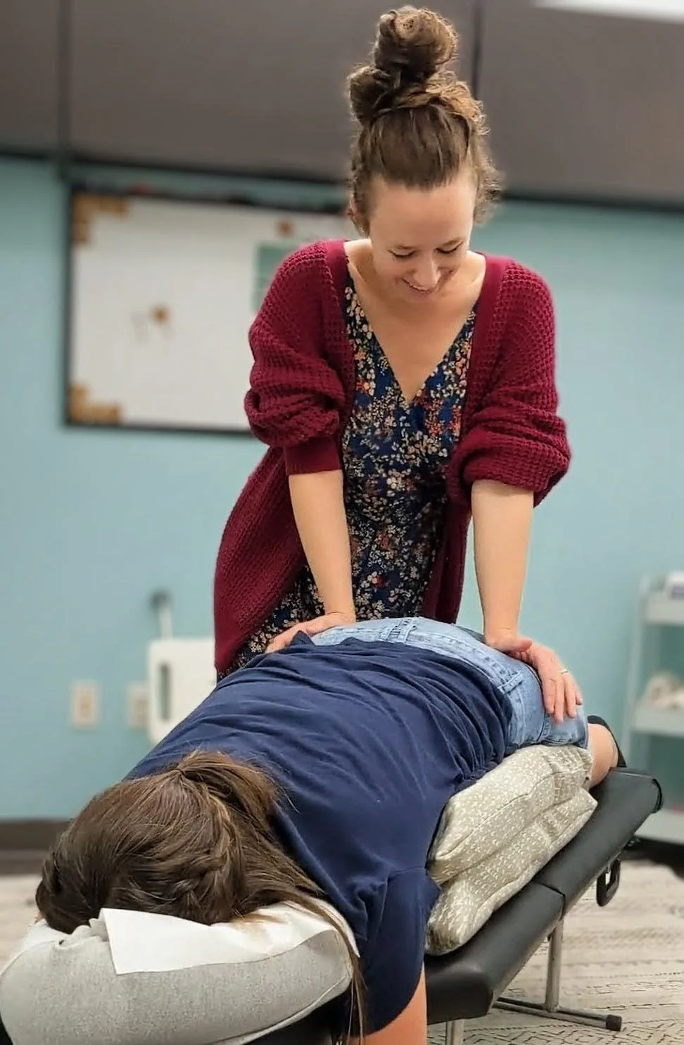 Woman giving a back massage to another person lying face down on a massage table in a room with teal walls.