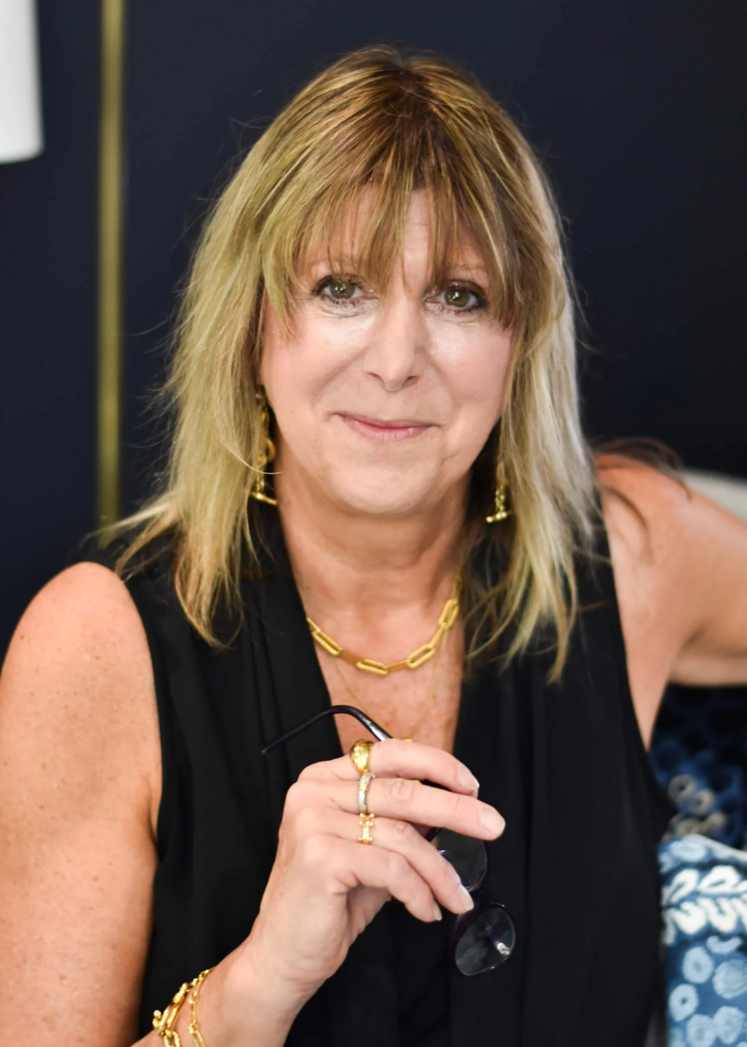 A woman with shoulder-length blonde hair, wearing a sleeveless black top, gold jewelry including a chain necklace, earrings, bracelets, and rings, holding glasses in her right hand, and looking at the camera with a slight smile.