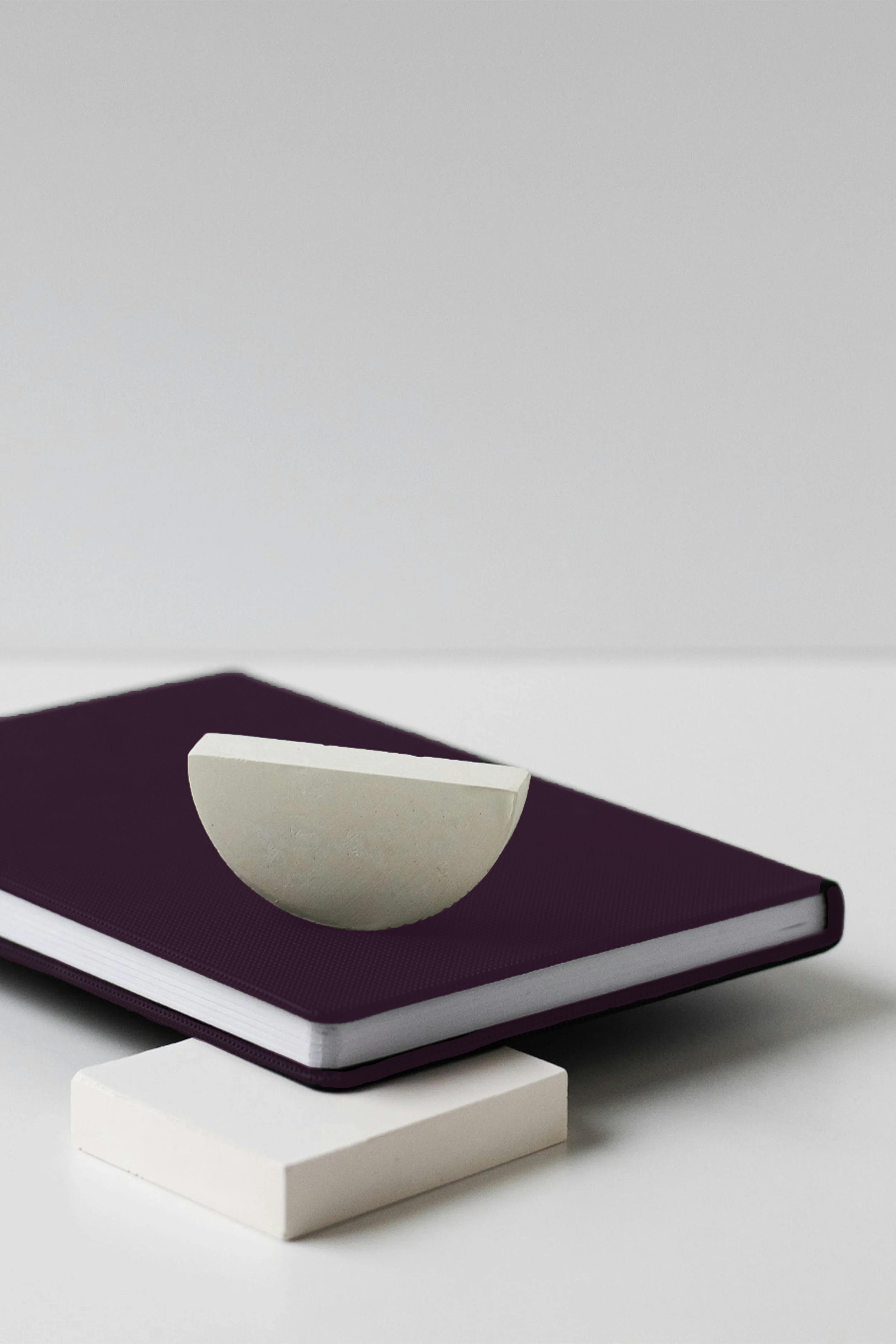 A minimalist still life composition with a closed purple notebook, a half circle white sculpture on top, and a rectangular white block underneath, all arranged on a plain white surface against a neutral background.