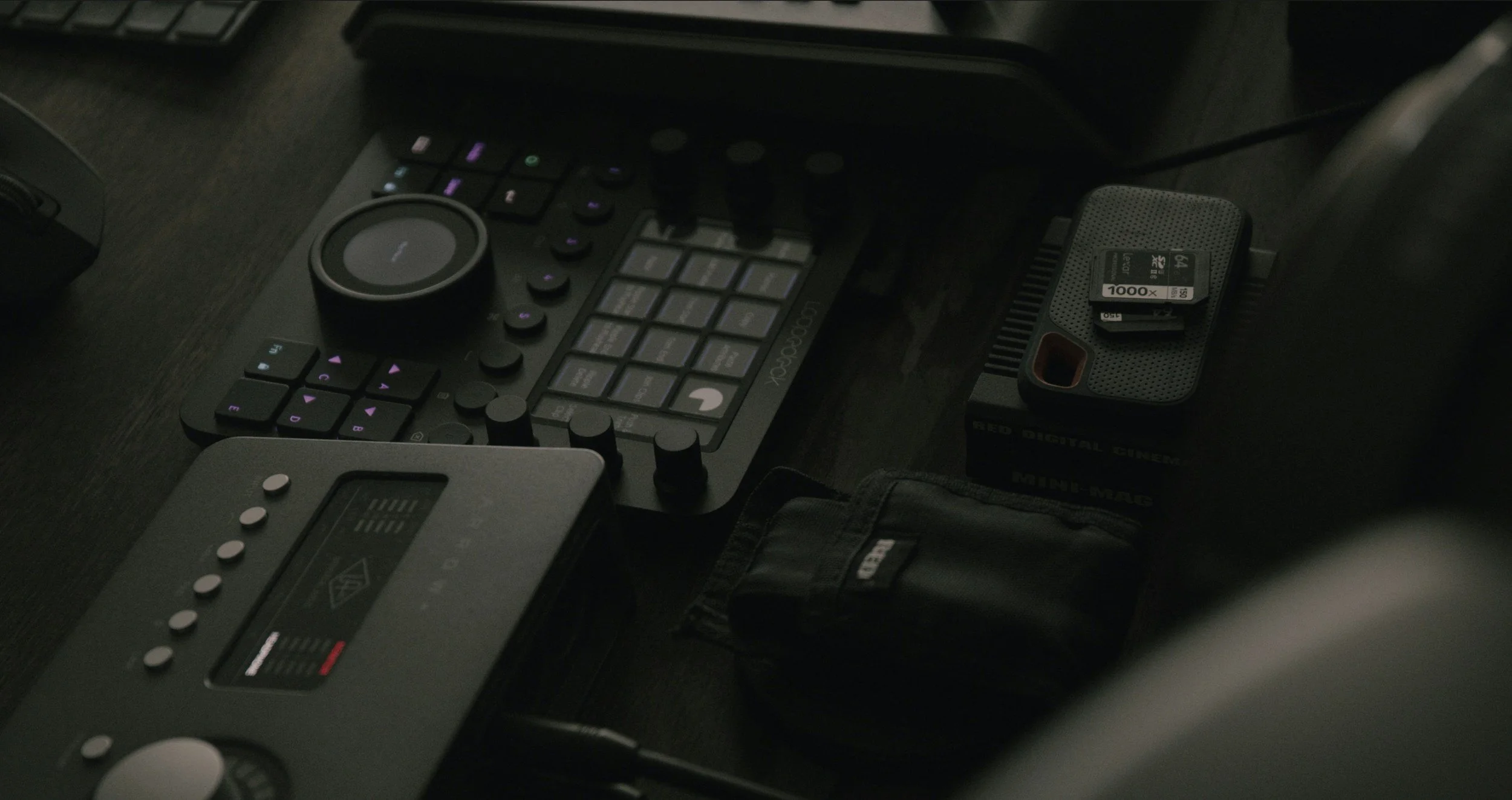 A dark workspace with a digital audio controller, a small black box with storage cards, and other electronic accessories on a black desk.